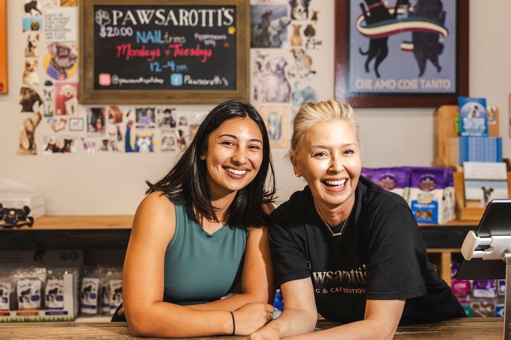 Two employees smile while working at a pet store called Pawsabotics, with pet supplies and decorative wall art visible in background.
