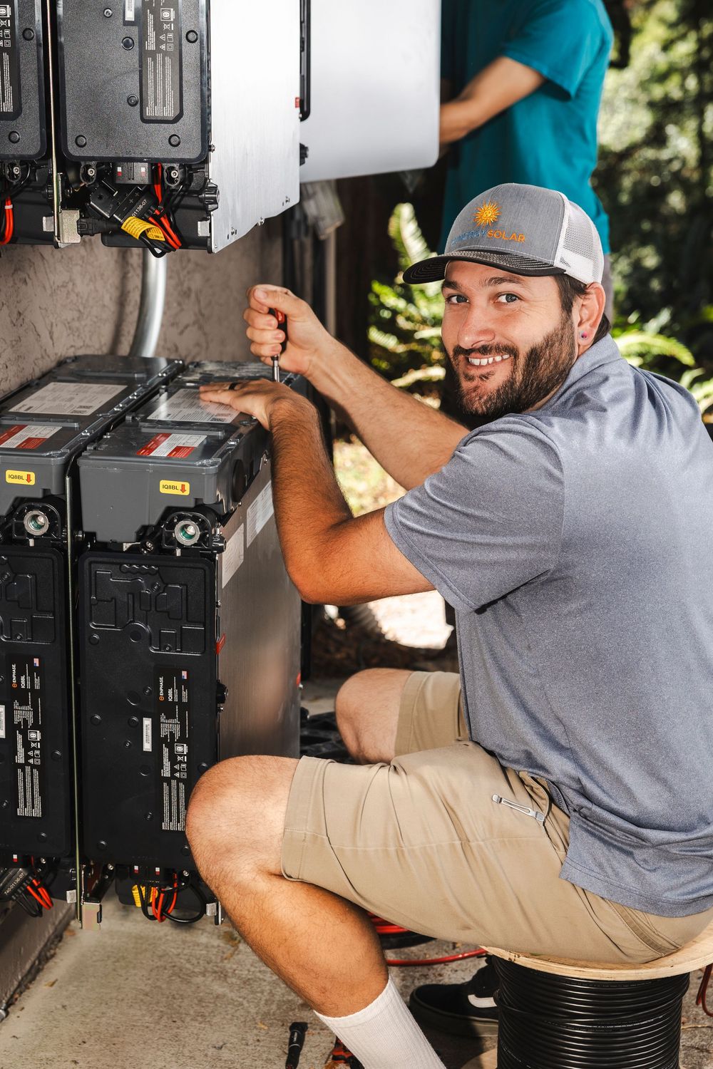 Smiling technician in gray hat and shirt works on electrical equipment while kneeling on outdoor service platform.