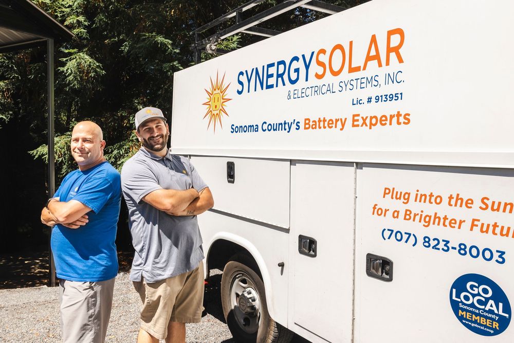 Two workers stand confidently in front of a Synergy Solar company van displaying their battery expertise services.