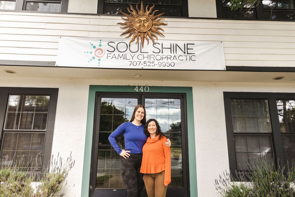 Two people stand smiling in front of Soul Shine Family Chiropractic office entrance with decorative sun sign above.