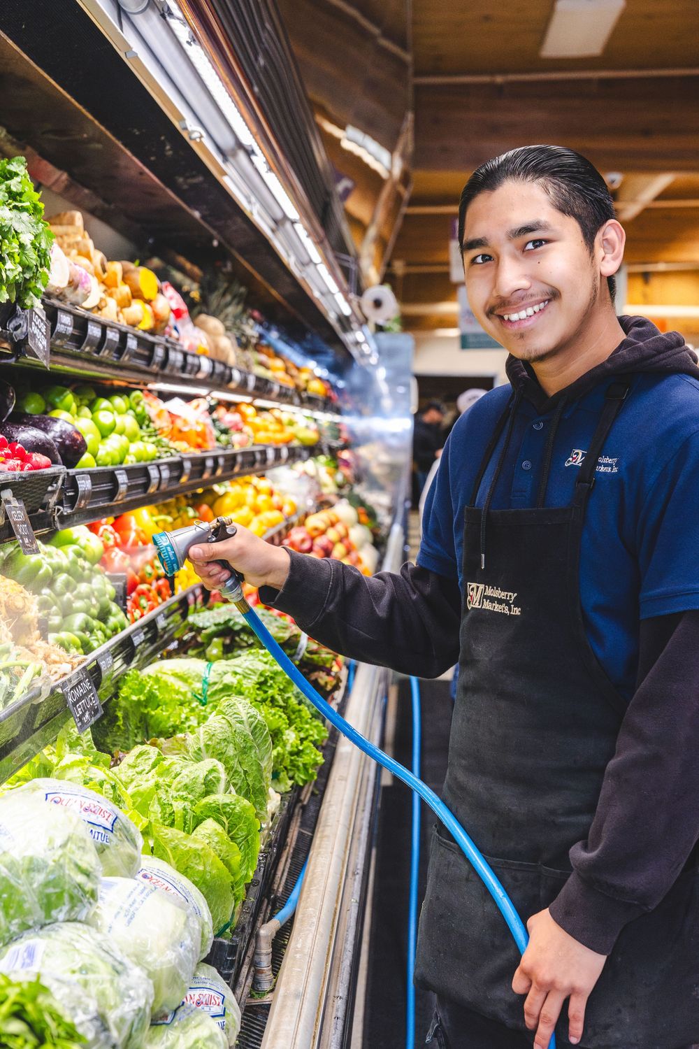 Grocery store worker in blue uniform arranges fresh produce in well-stocked display case at supermarket.