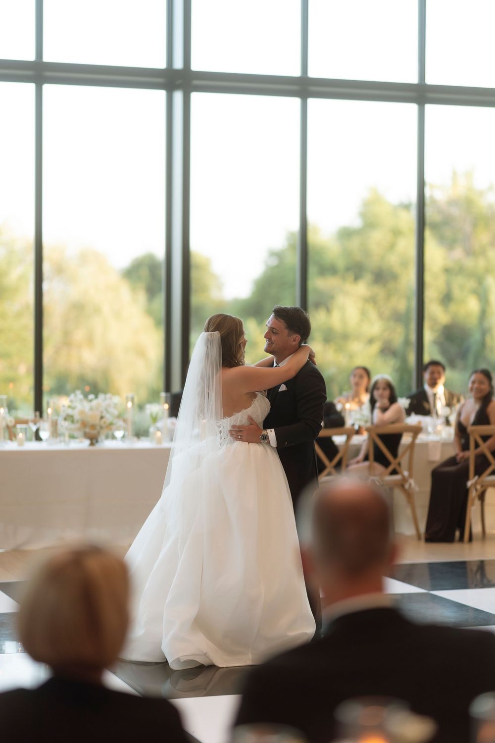 A couple shares their first dance at a wedding reception in a bright, window-lined venue with guests watching.