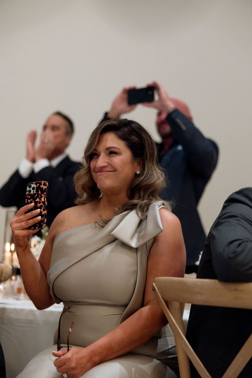 Guest in a taupe one-shoulder dress smiles while holding a phone at a formal event.
