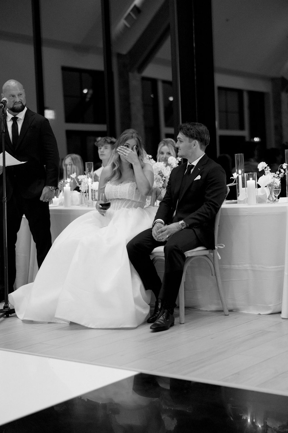 A bride in a white gown and groom in a black suit sit together at their wedding reception.