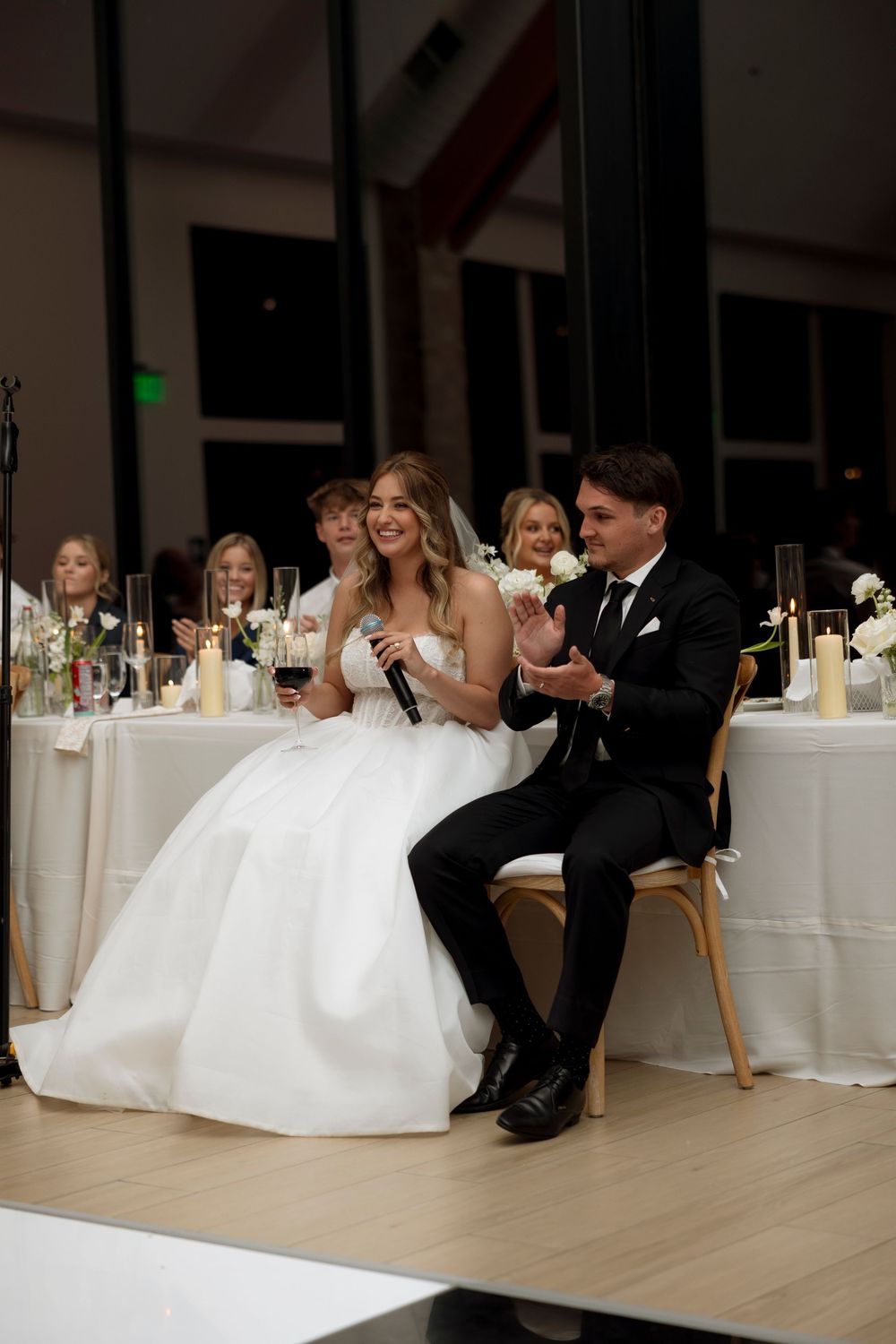 Wedding guests seated at elegant white-clothed tables enjoy a reception in a modern venue with light wood floors.