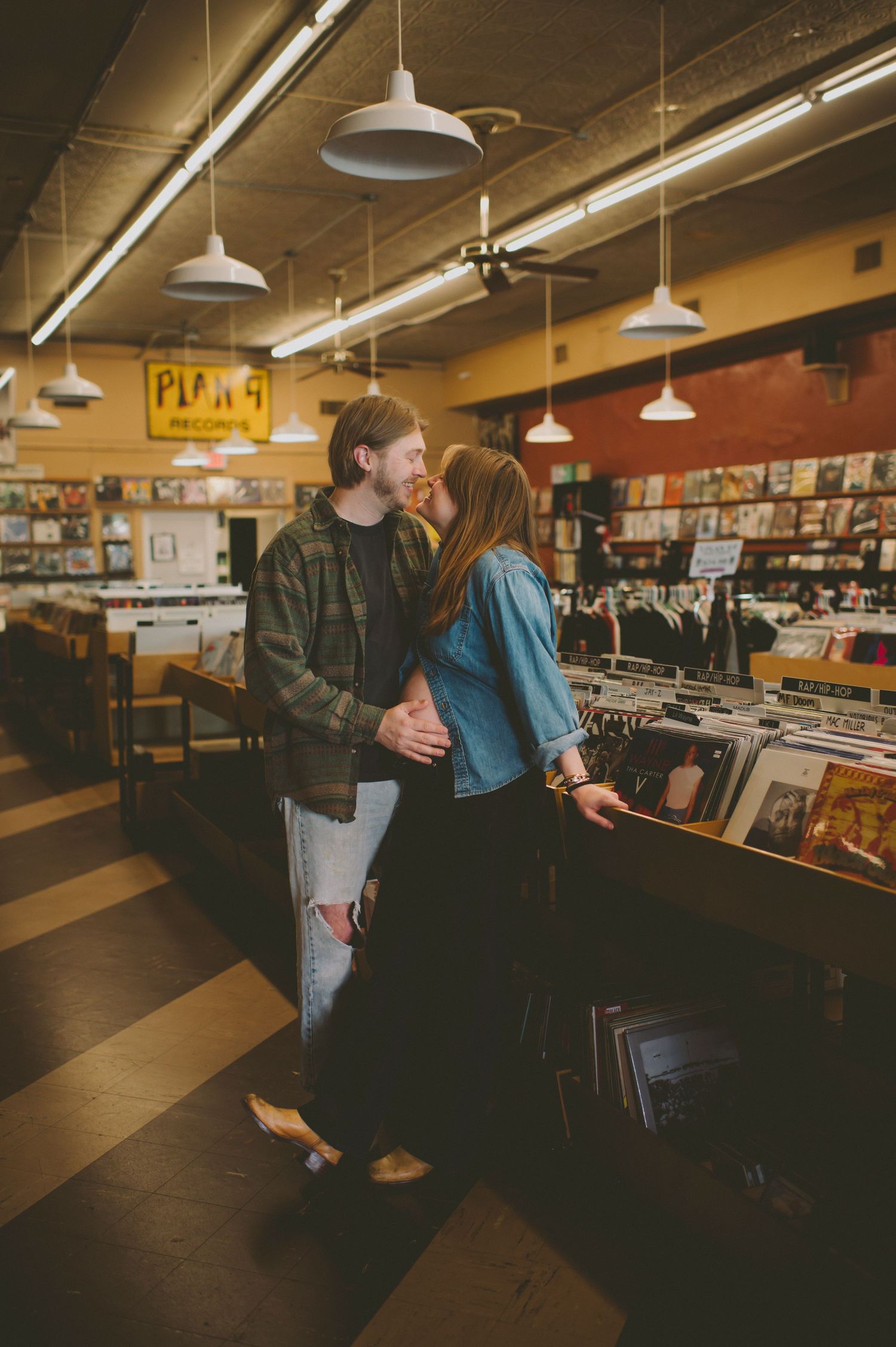 A couple browses vinyl records together in a dimly lit record store with vintage pendant lighting overhead.
