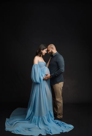 A couple in a romantic maternity photoshoot poses against a dark background with the woman wearing a flowing blue gown.