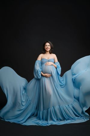 A flowing light blue maternity gown creates an ethereal silhouette against a dark backdrop in a studio portrait series.