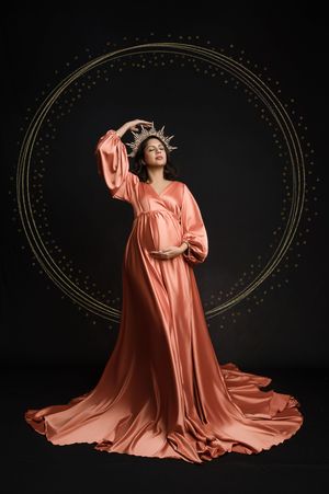 A figure in a flowing coral silk gown poses dramatically against a dark background with an illuminated circular frame.