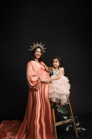 Studio portrait with two figures in elegant dresses against black background, one wearing a crown and coral gown.