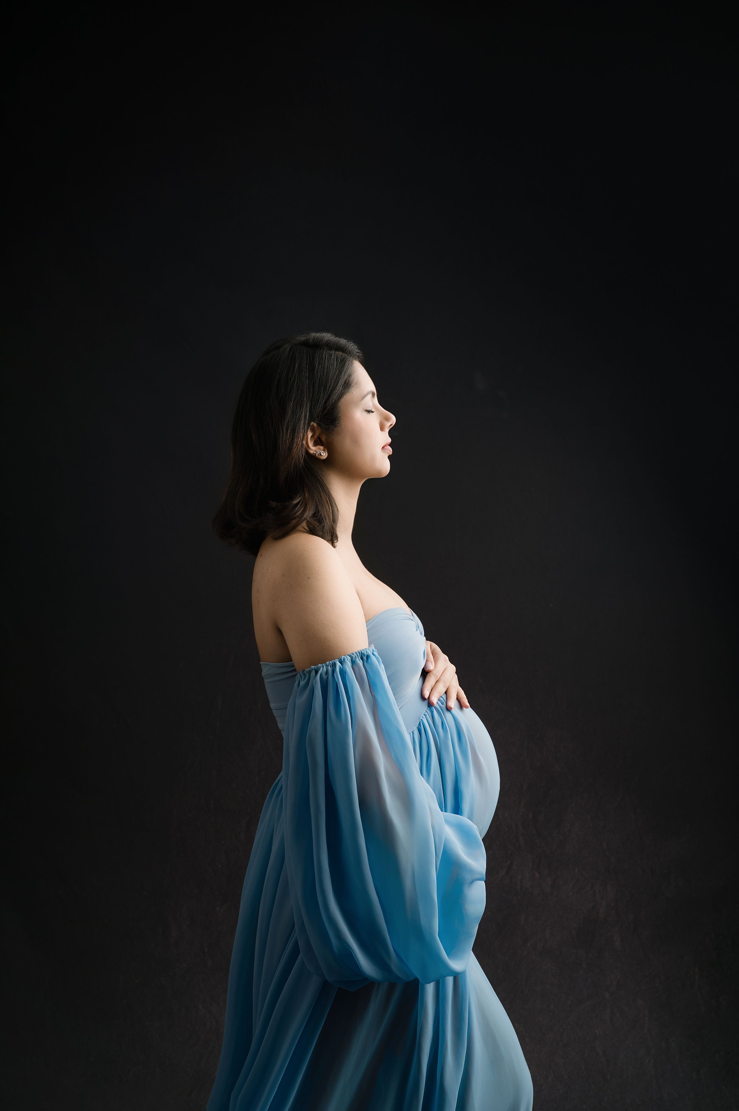 Figure in flowing light blue dress poses dramatically against dark background in artistic maternity photoshoot.