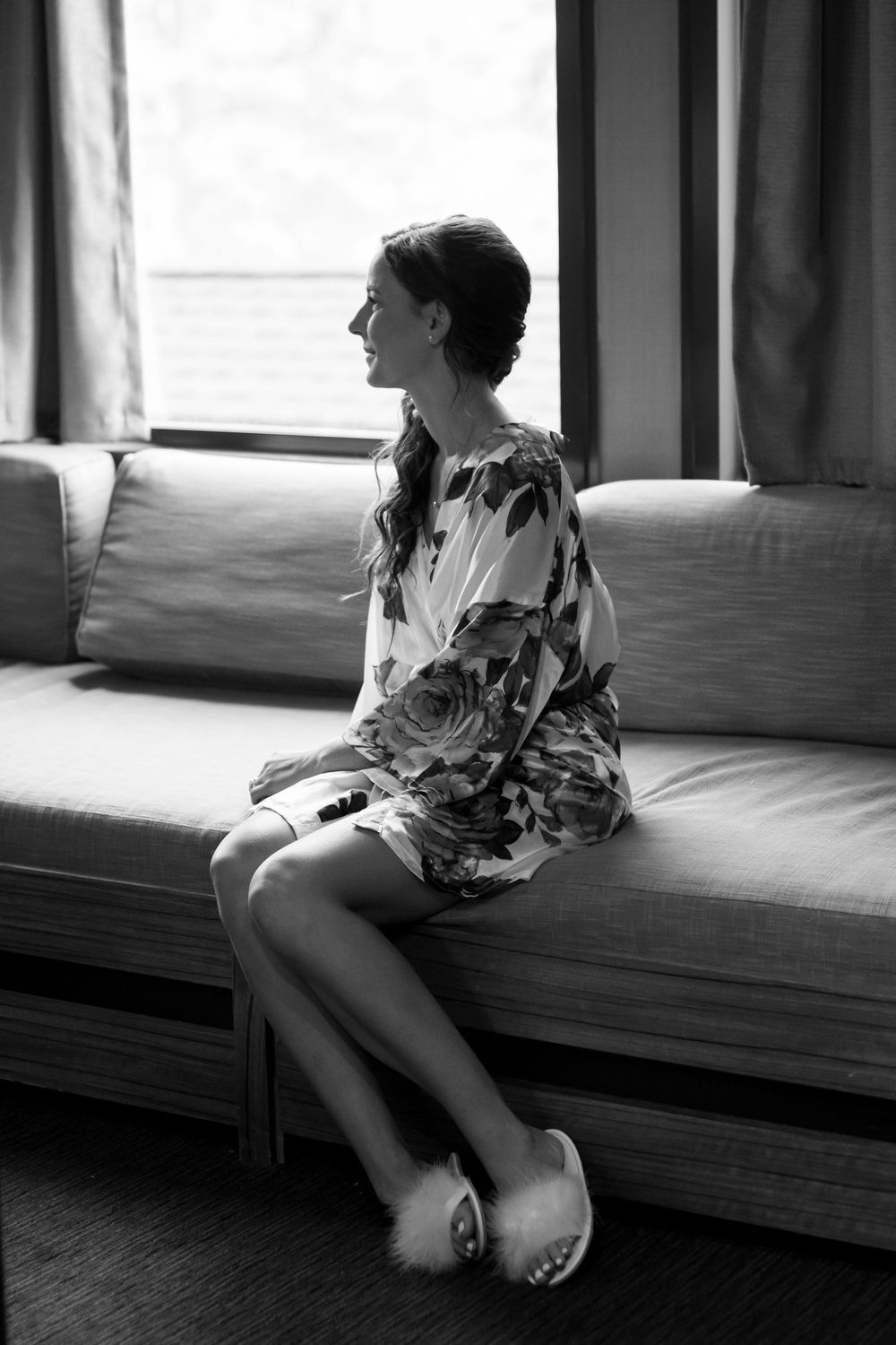 A silhouetted figure in floral dress sits on couch by window in black and white photograph.