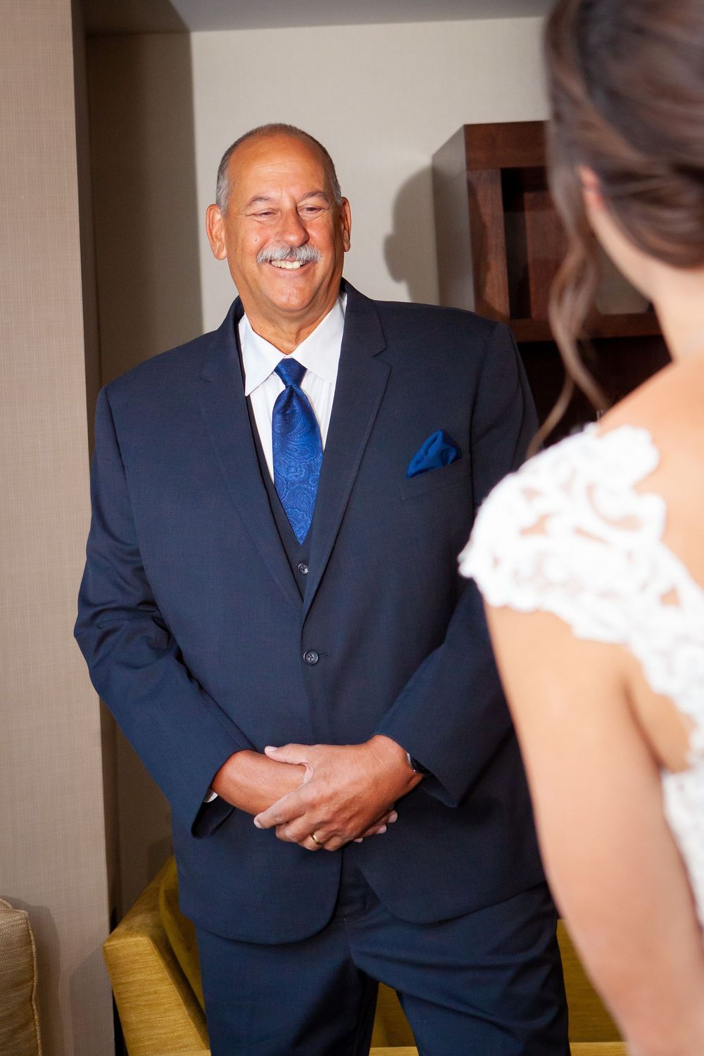 A man in a navy suit and blue tie smiles joyfully while looking at someone in a wedding dress.