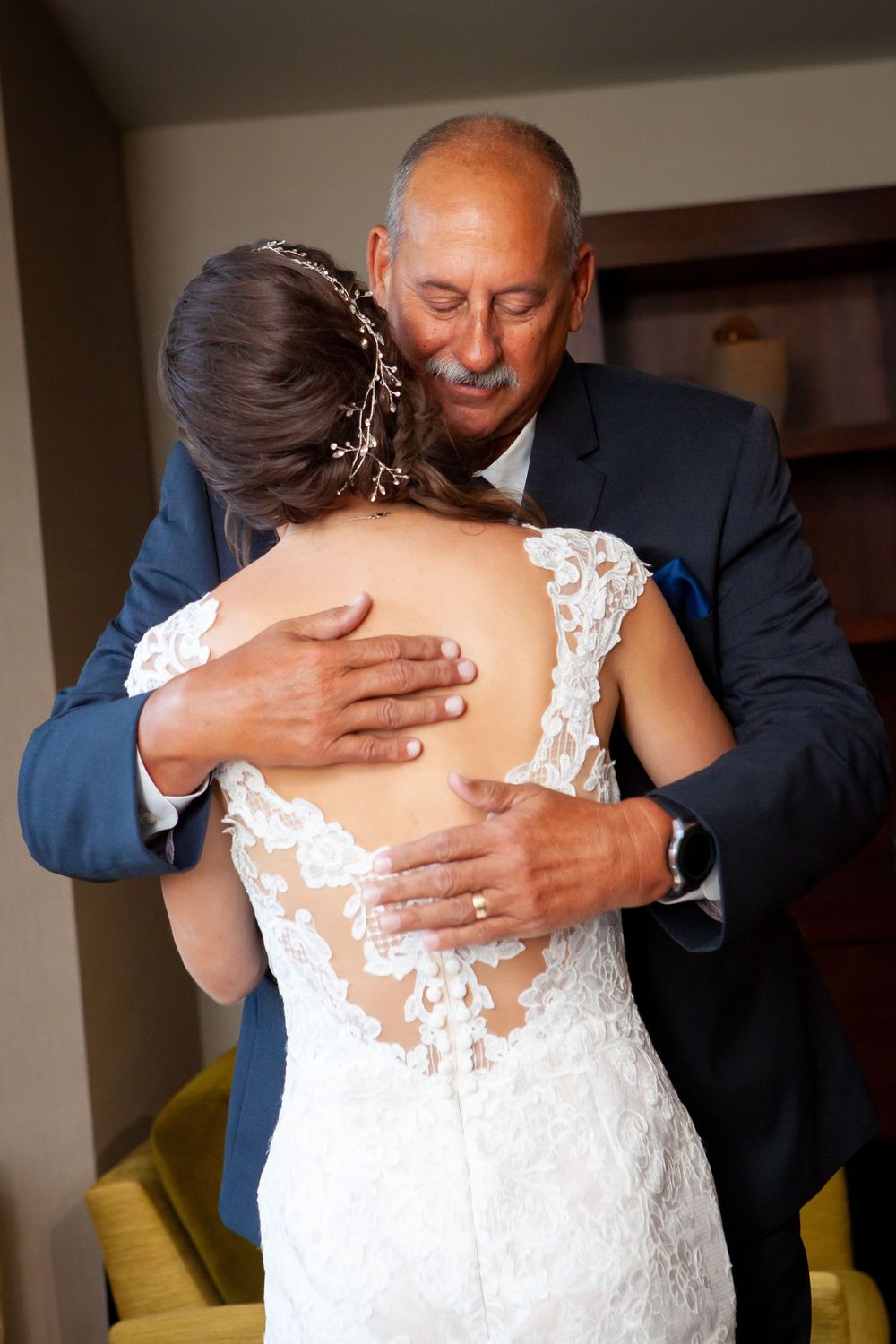 A tender moment between a bride in a lace wedding dress and an older man in a navy suit sharing an emotional embrace.