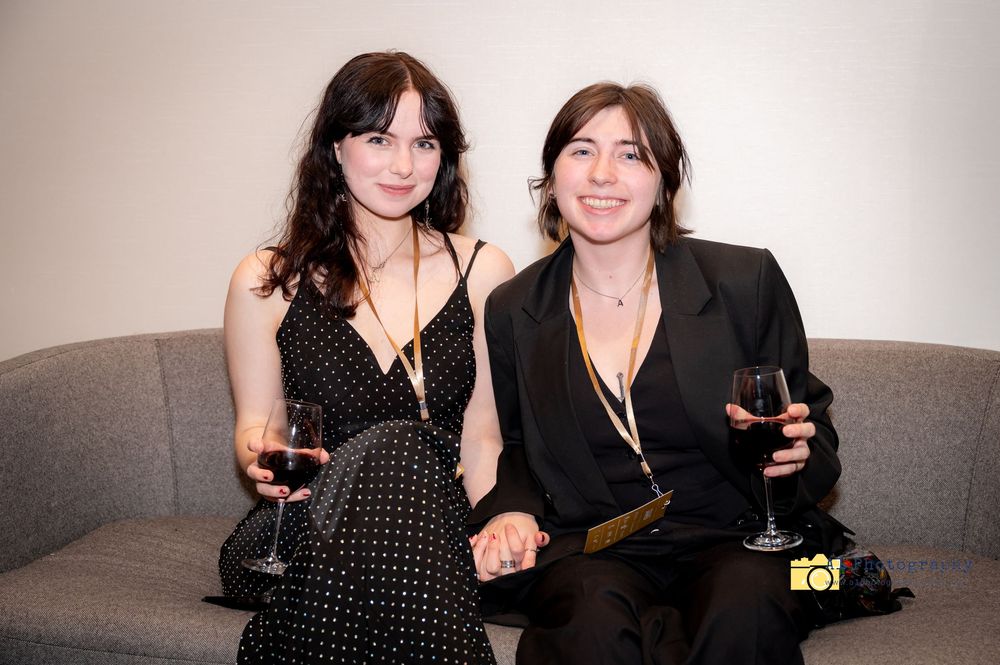Two people in black attire sit close together on a couch while enjoying glasses of red wine.