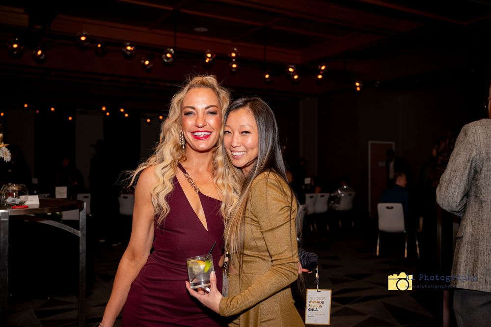 Two women smile together at an evening social event, one wearing a burgundy dress and one in a tan jacket.