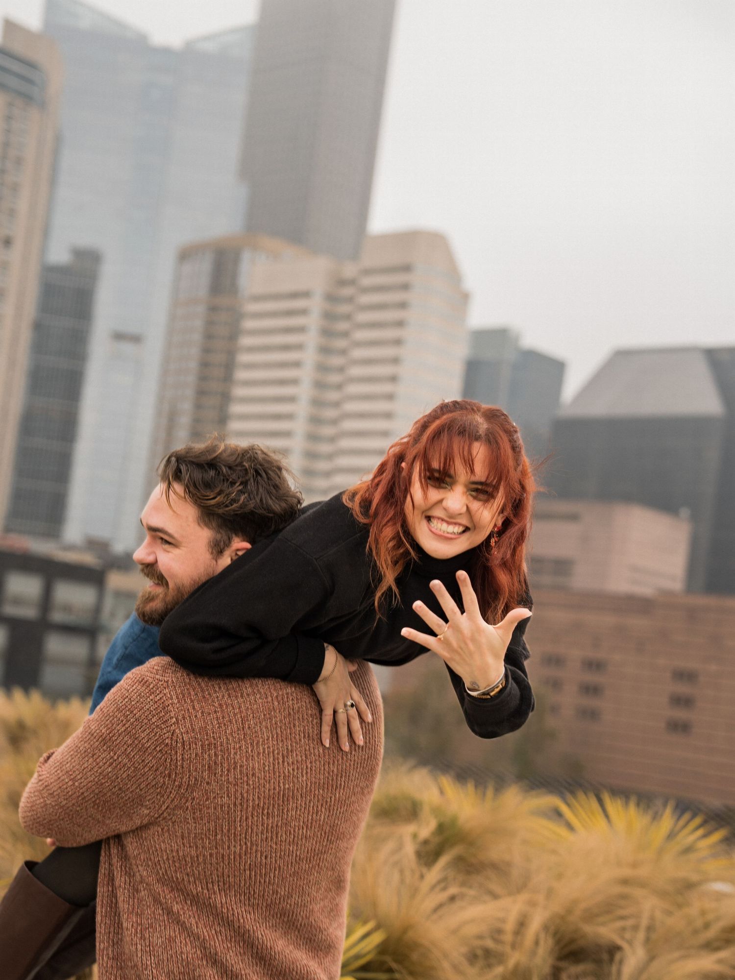 A couple embraces while sitting on concrete steps outside a modern high-rise building in warm sunlight.