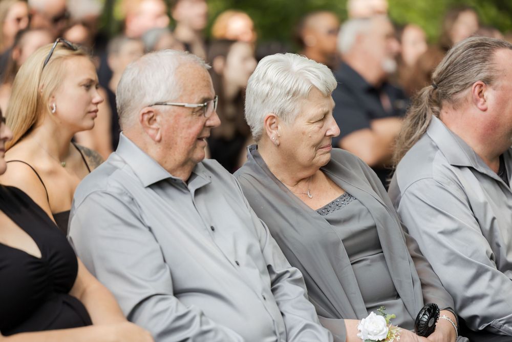 Wedding guests seated outdoors in gray and black attire smile during a ceremony.