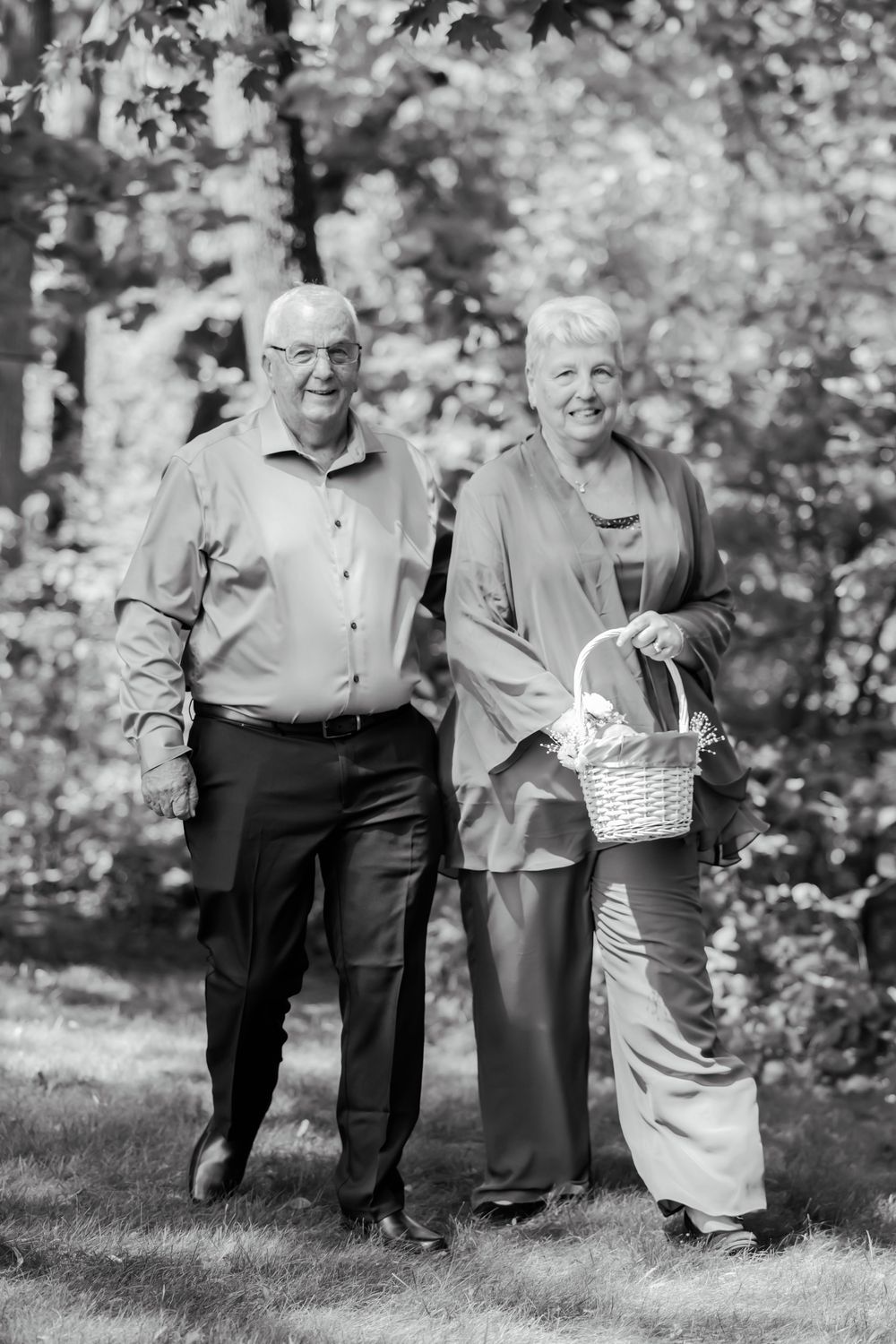 An elderly couple walks together through a wooded area, with the woman carrying a basket.
