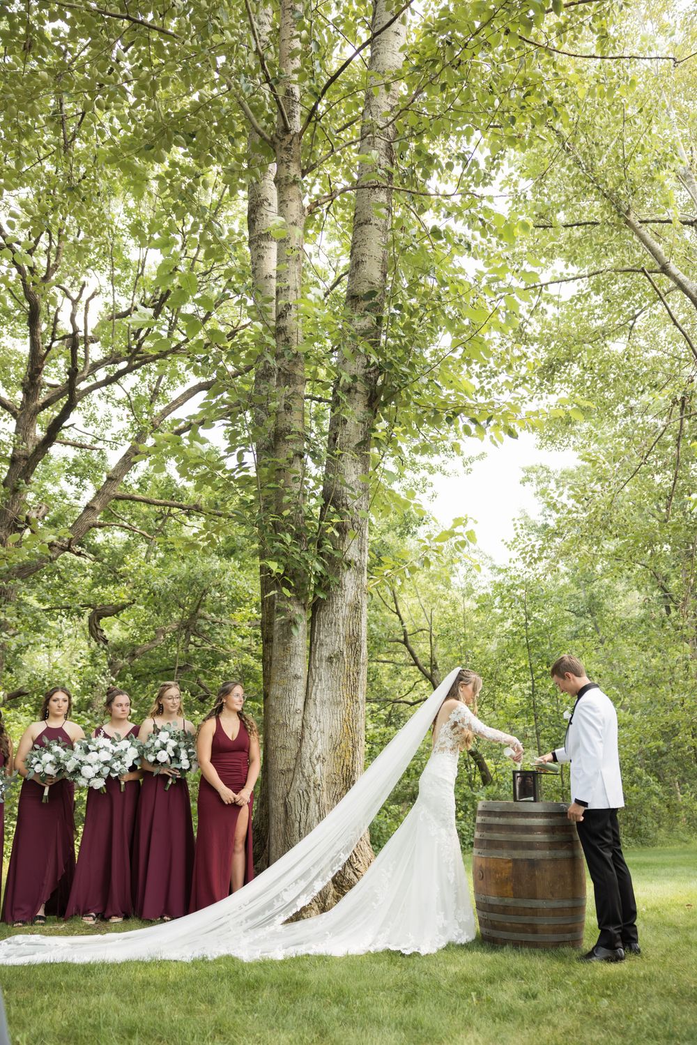 Bridal party in burgundy dresses stands in wooded outdoor setting with wine barrels.