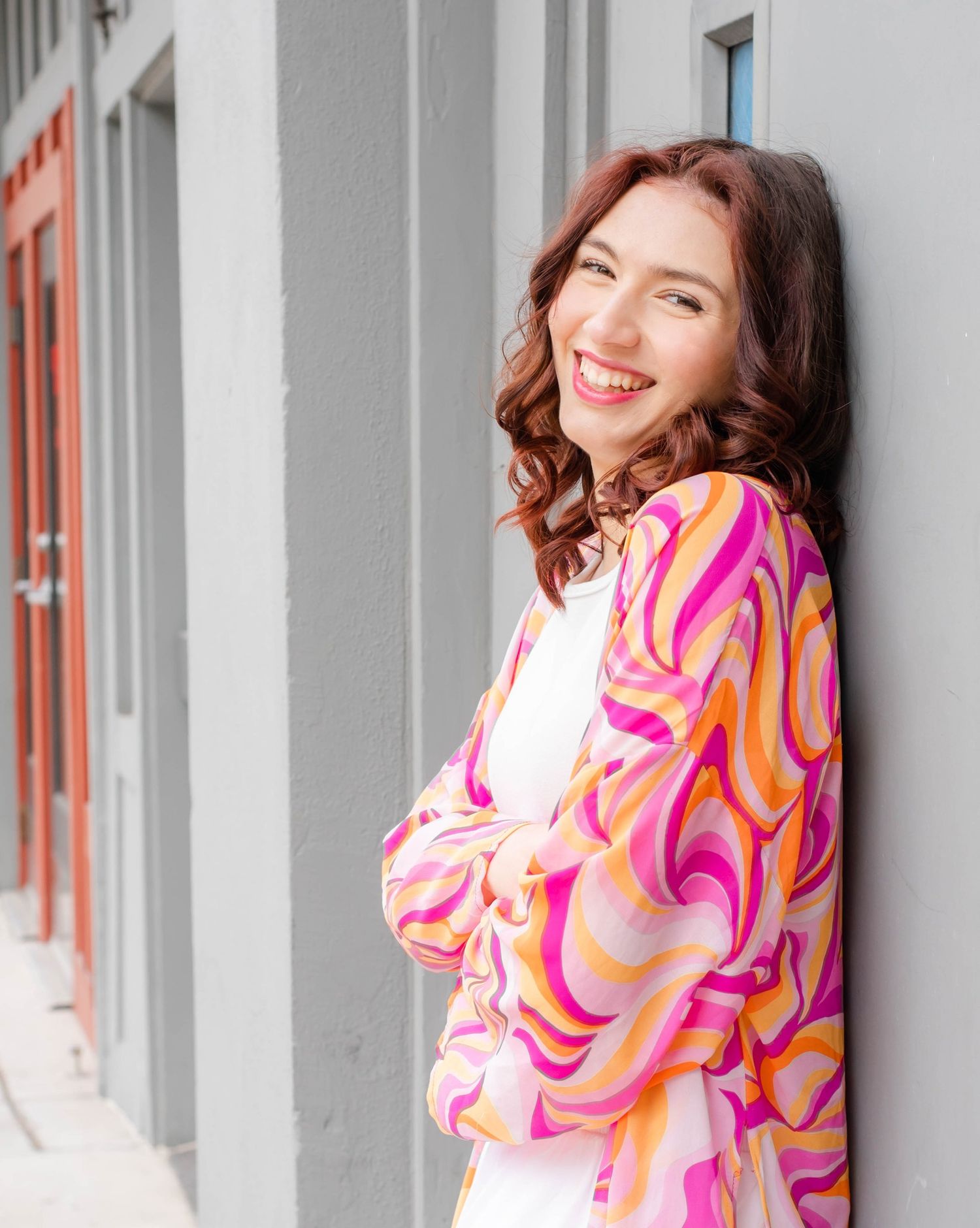 Woman in vibrant pink and orange swirled pattern dress poses against a light gray wall with windows.