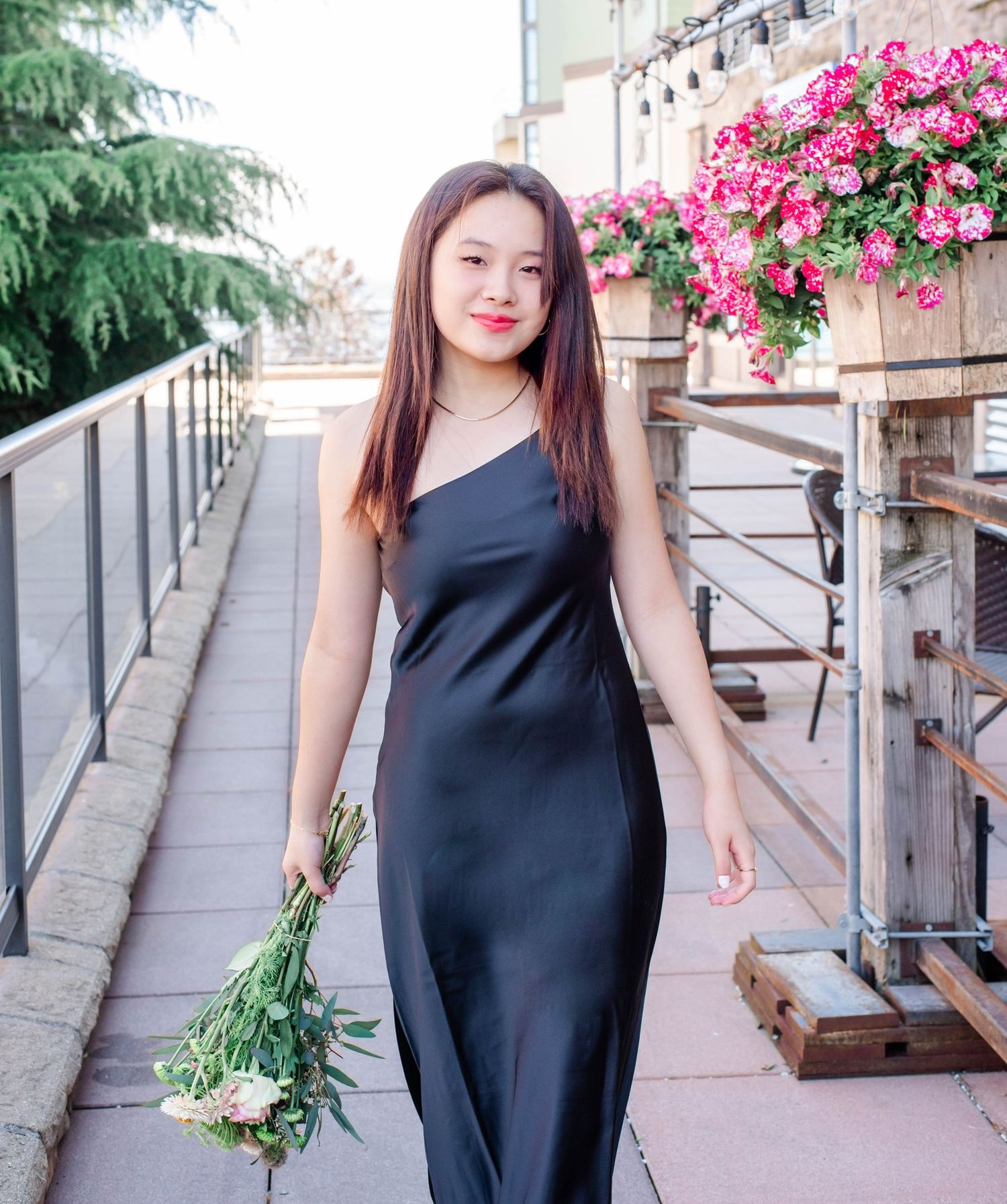 Elegant black dress against pink flowers and greenery on an outdoor walkway with railing.