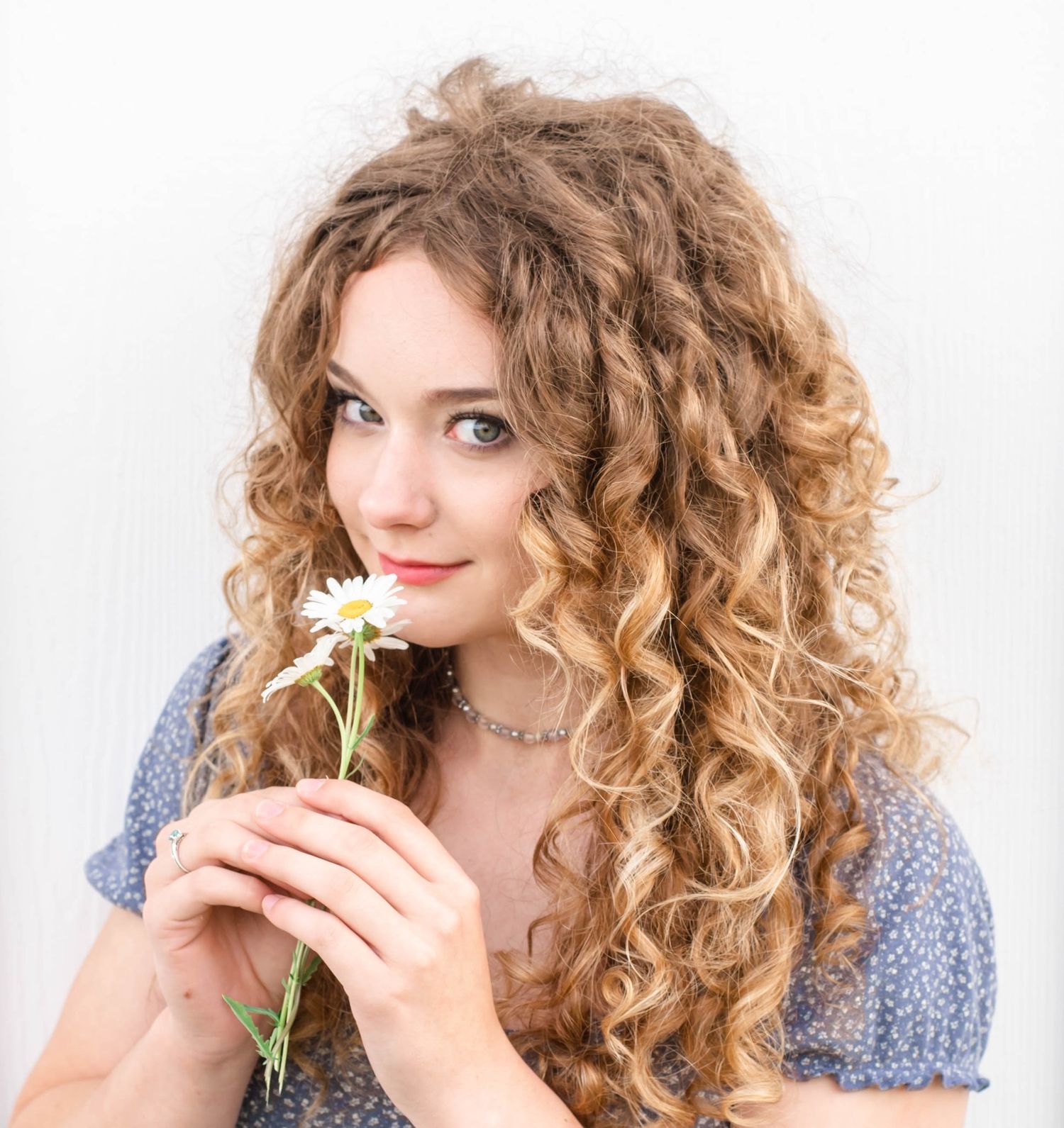 Person with curly blonde hair in blue floral dress poses with white flower in creative portrait series.