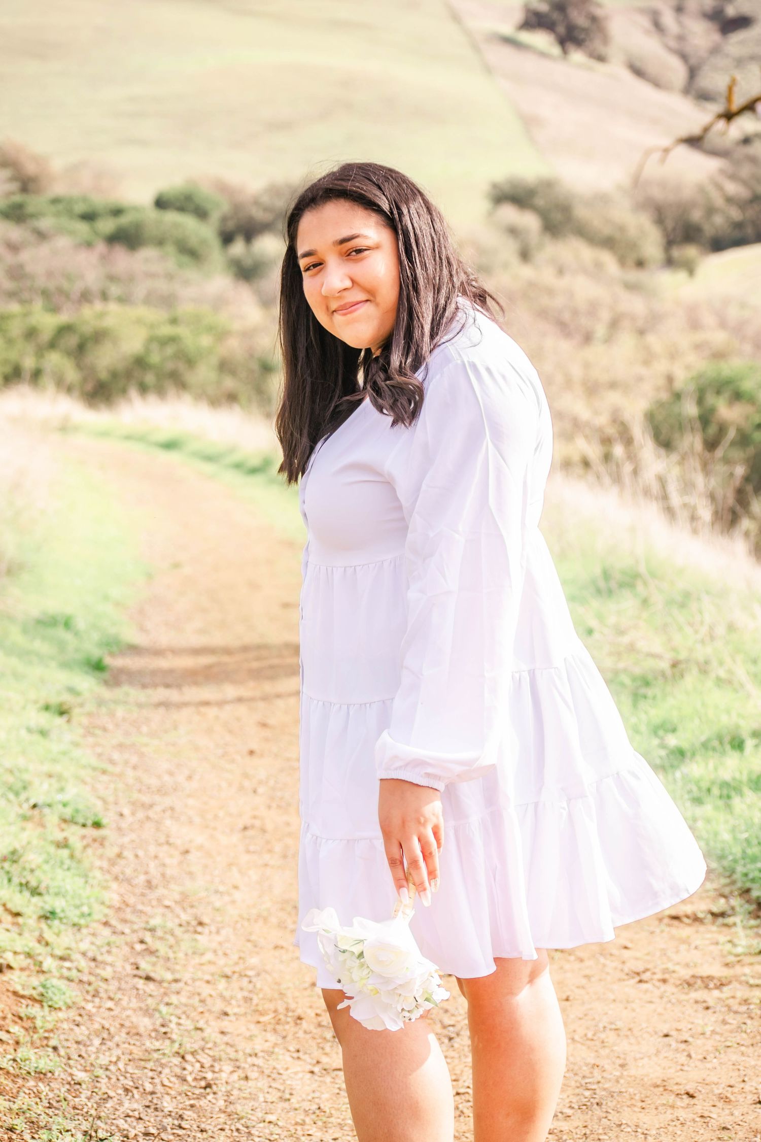 A person in a flowy white dress poses on a dirt trail against a golden hillside backdrop in warm sunlight.