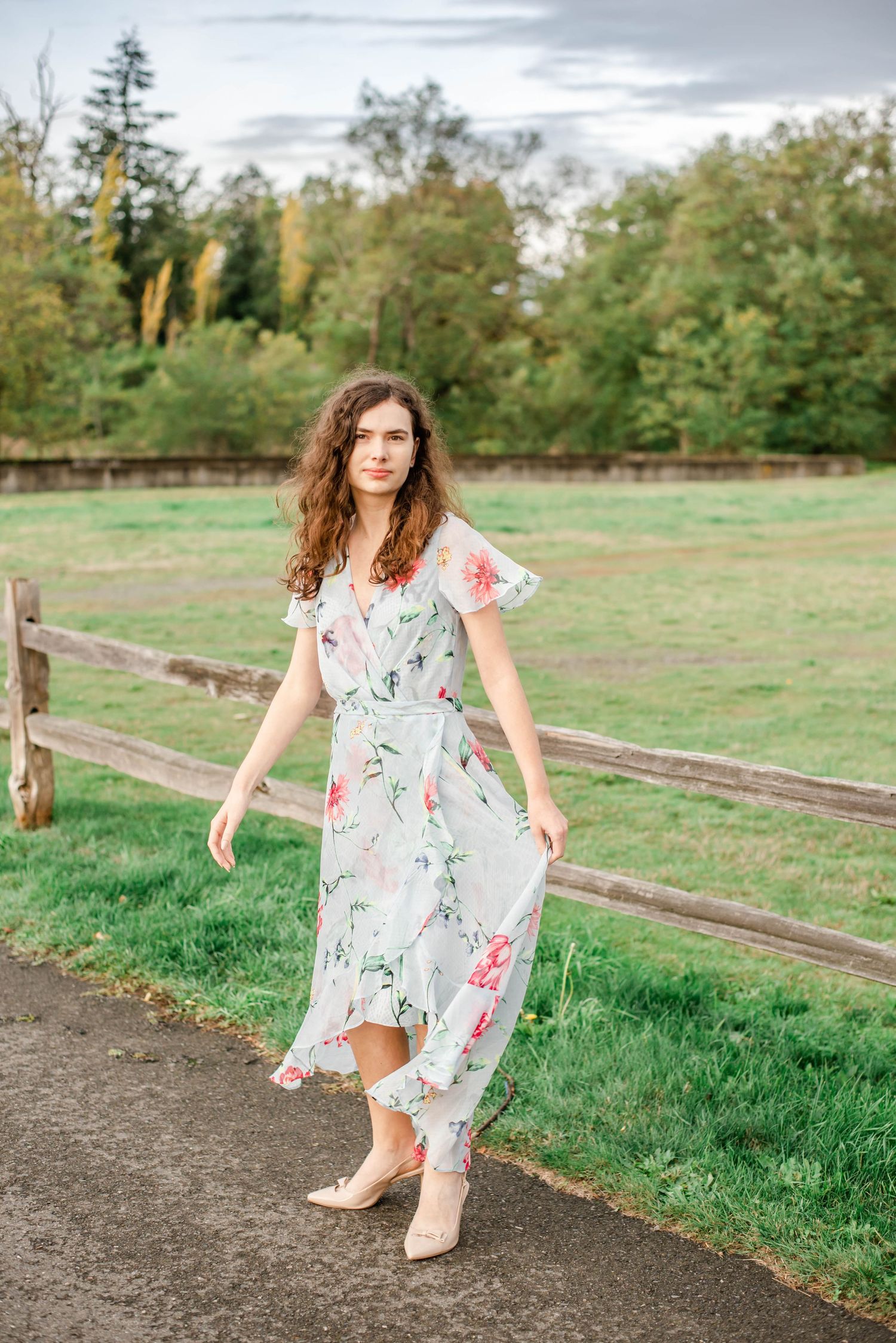 A person in a floral white dress twirls playfully on a dirt path next to a wooden fence and green field.