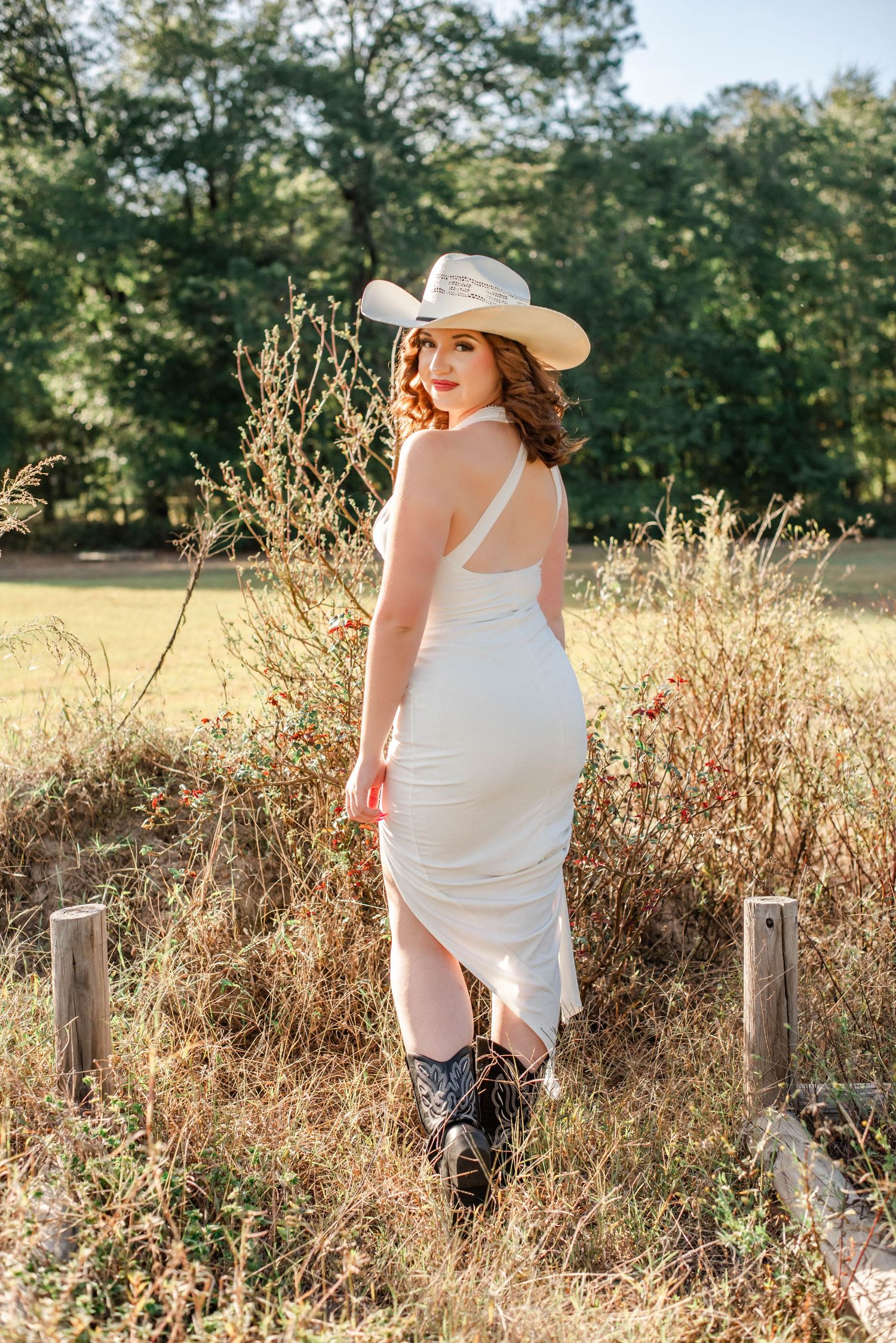 A person in a white dress and sun hat poses gracefully against a rustic wooden fence in a sunlit meadow.