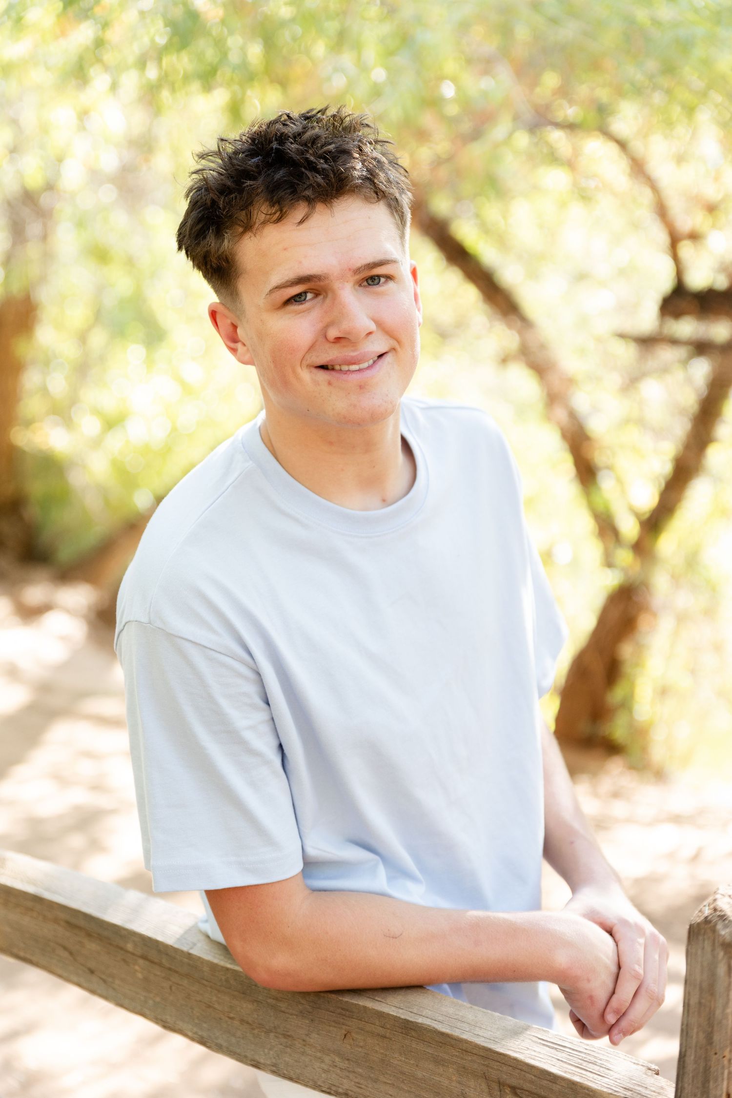 A senior boy in a light colored shirt leans against a wood fence smiling at the camera southern utah
