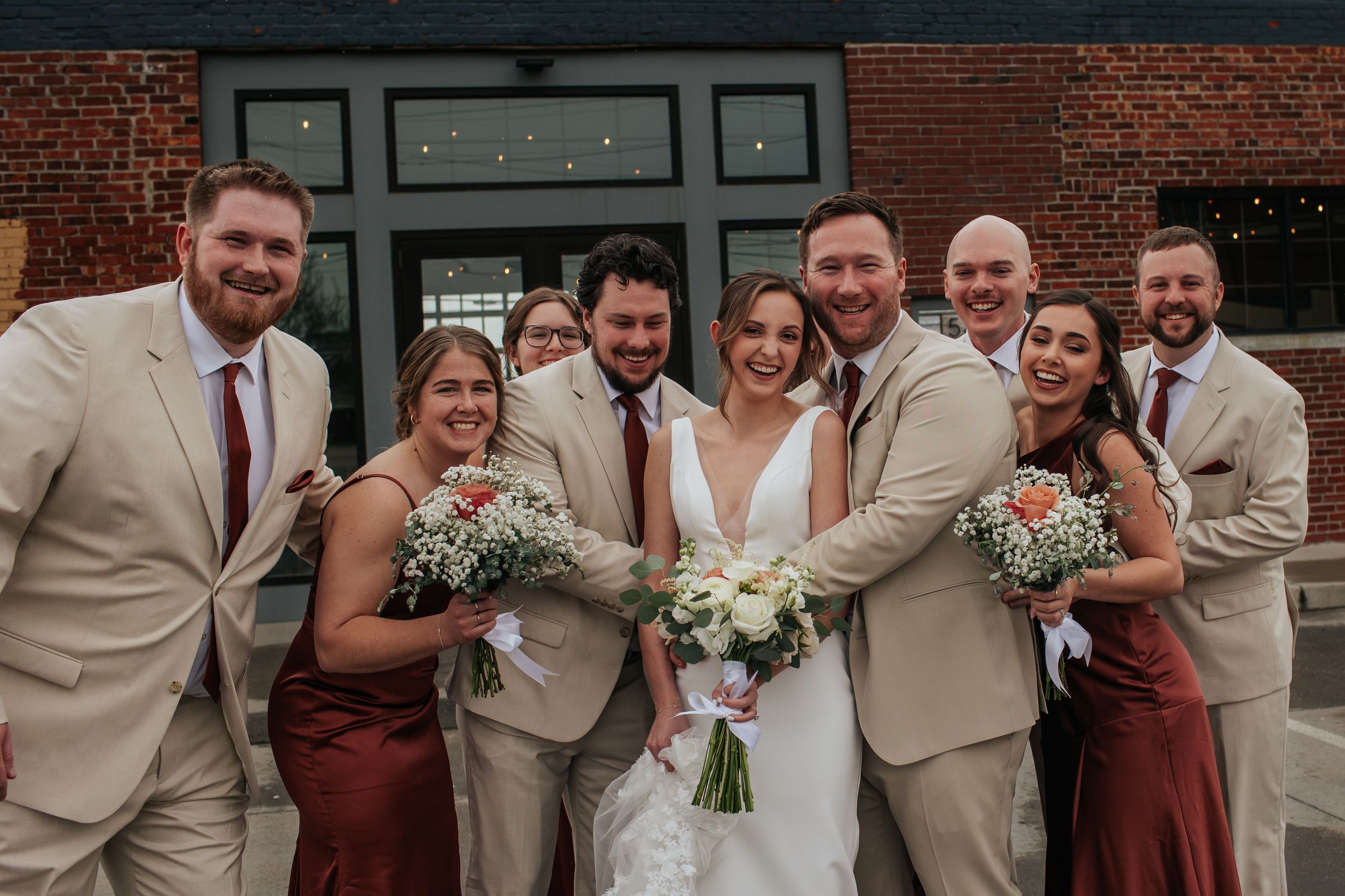 A wedding party poses together wearing burgundy and beige attire while holding floral bouquets outside a brick building.