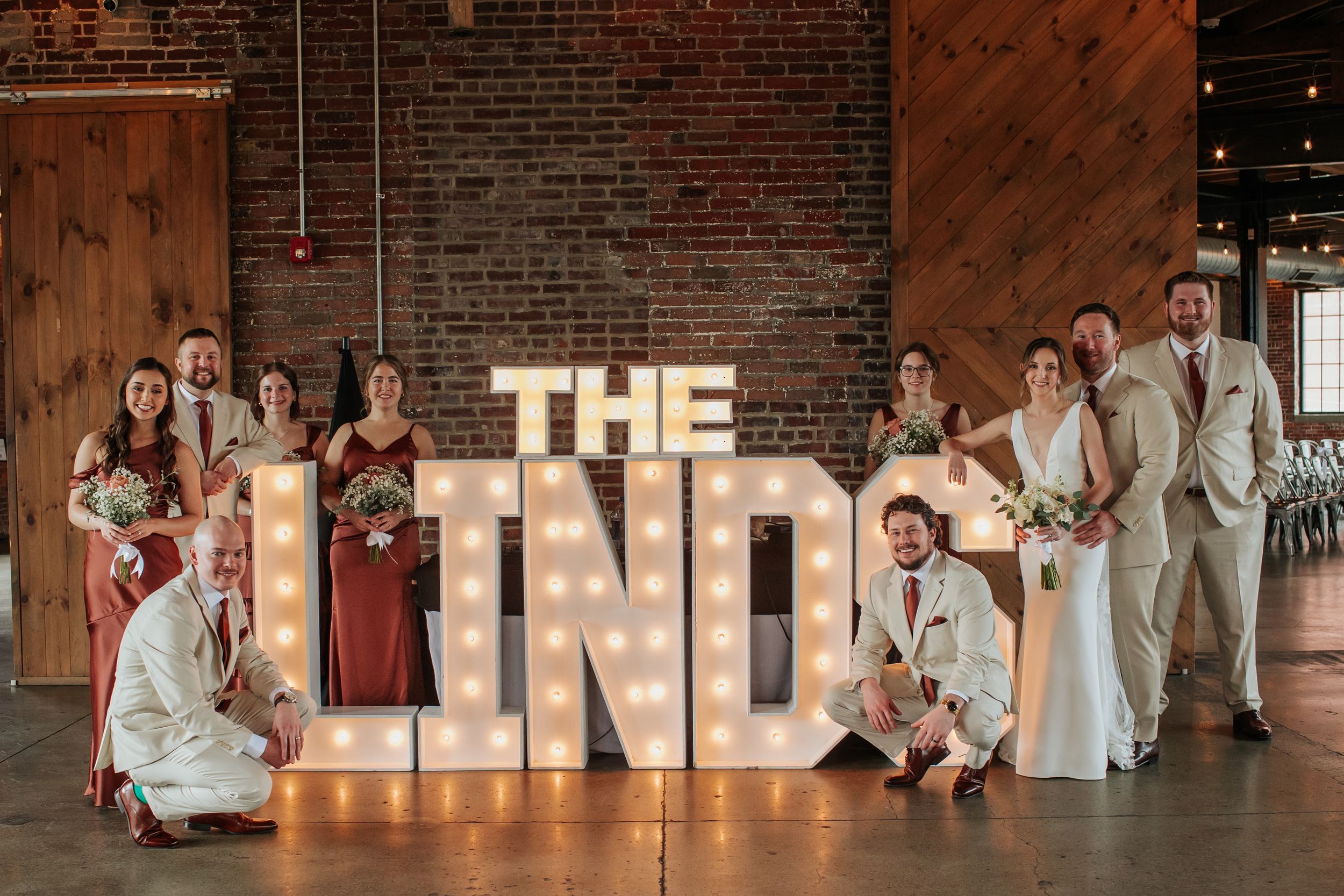 Wedding party poses in rustic brick venue with illuminated marquee letters creating a warm, romantic atmosphere.