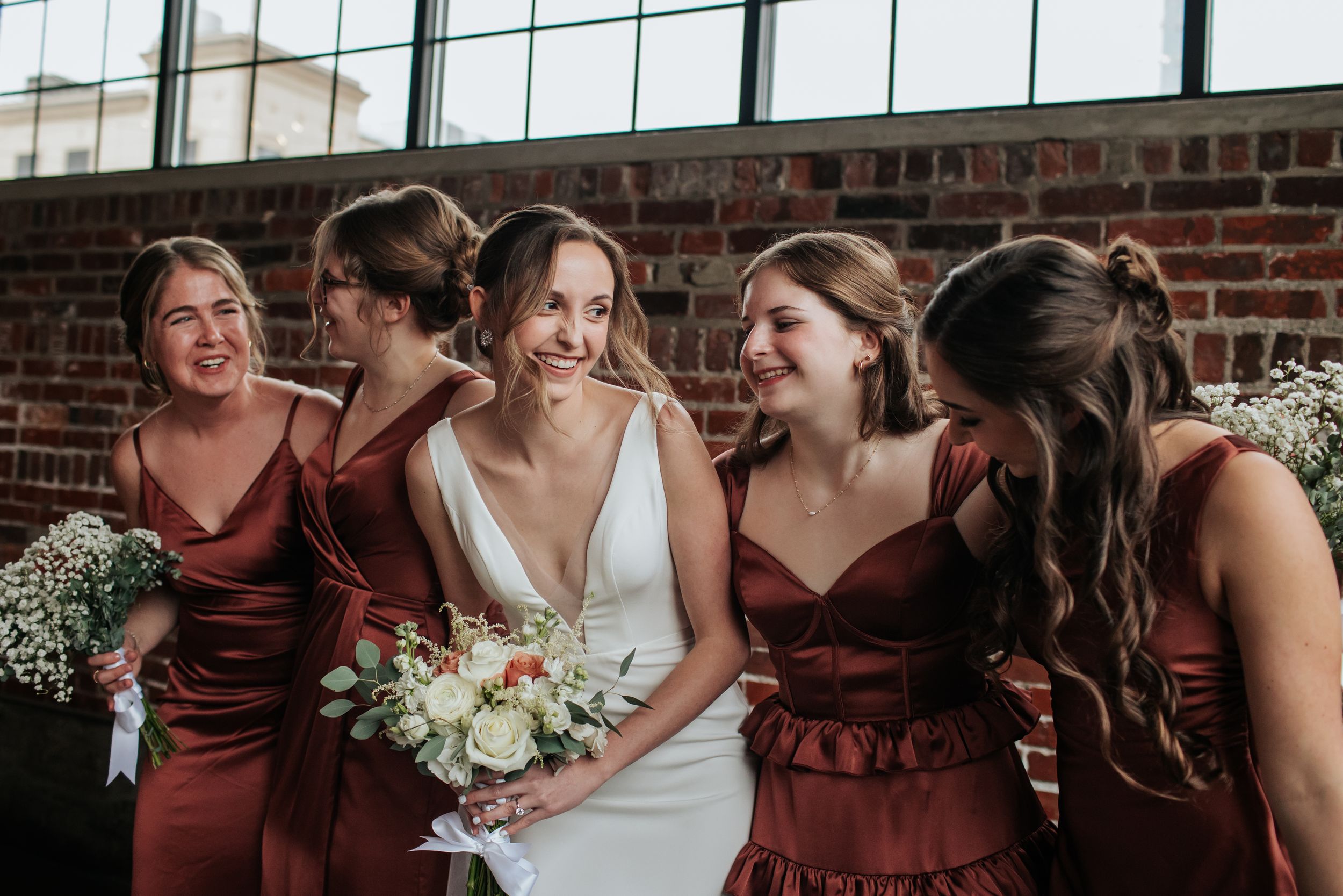 A wedding party in burgundy and white dresses poses together in front of a brick wall.