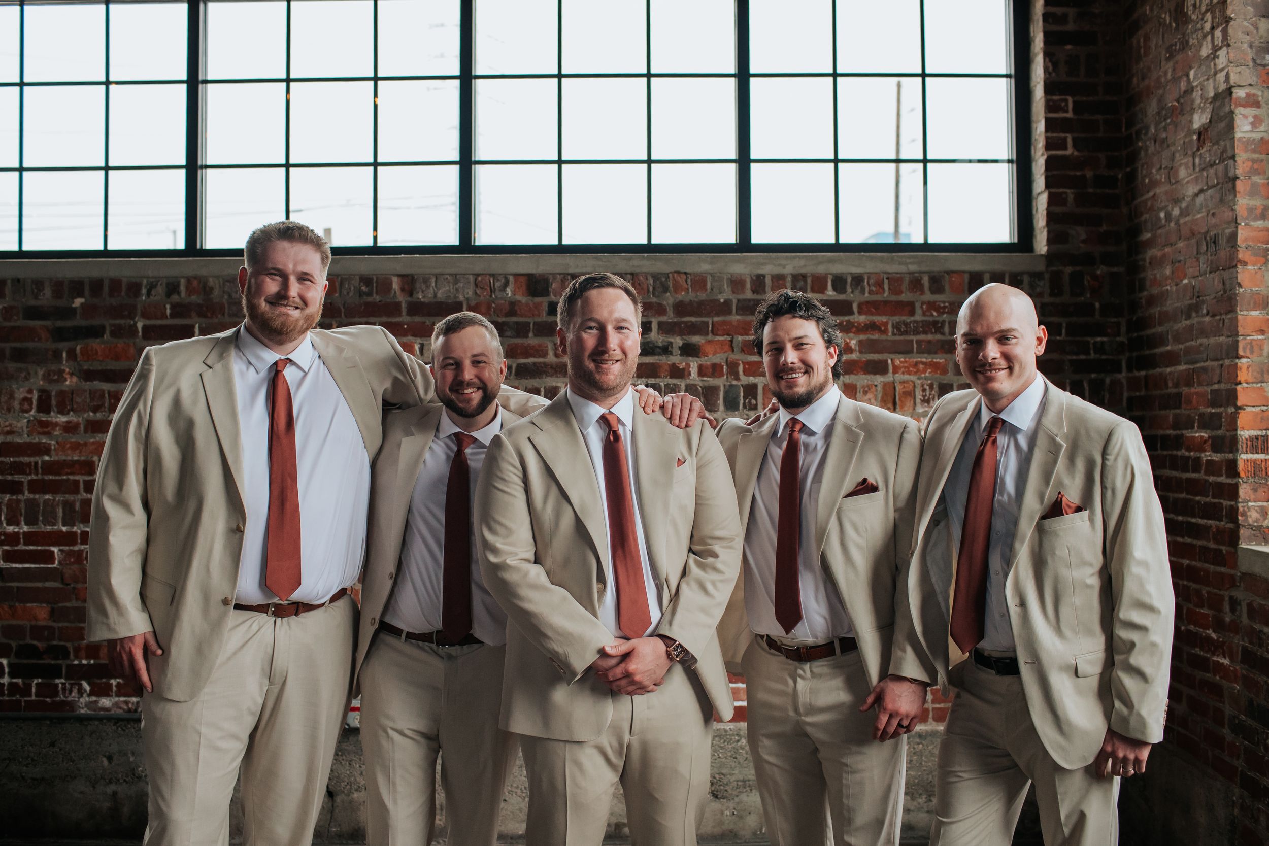 Groomsmen in beige suits with red ties pose together against a brick wall in an industrial-style venue.