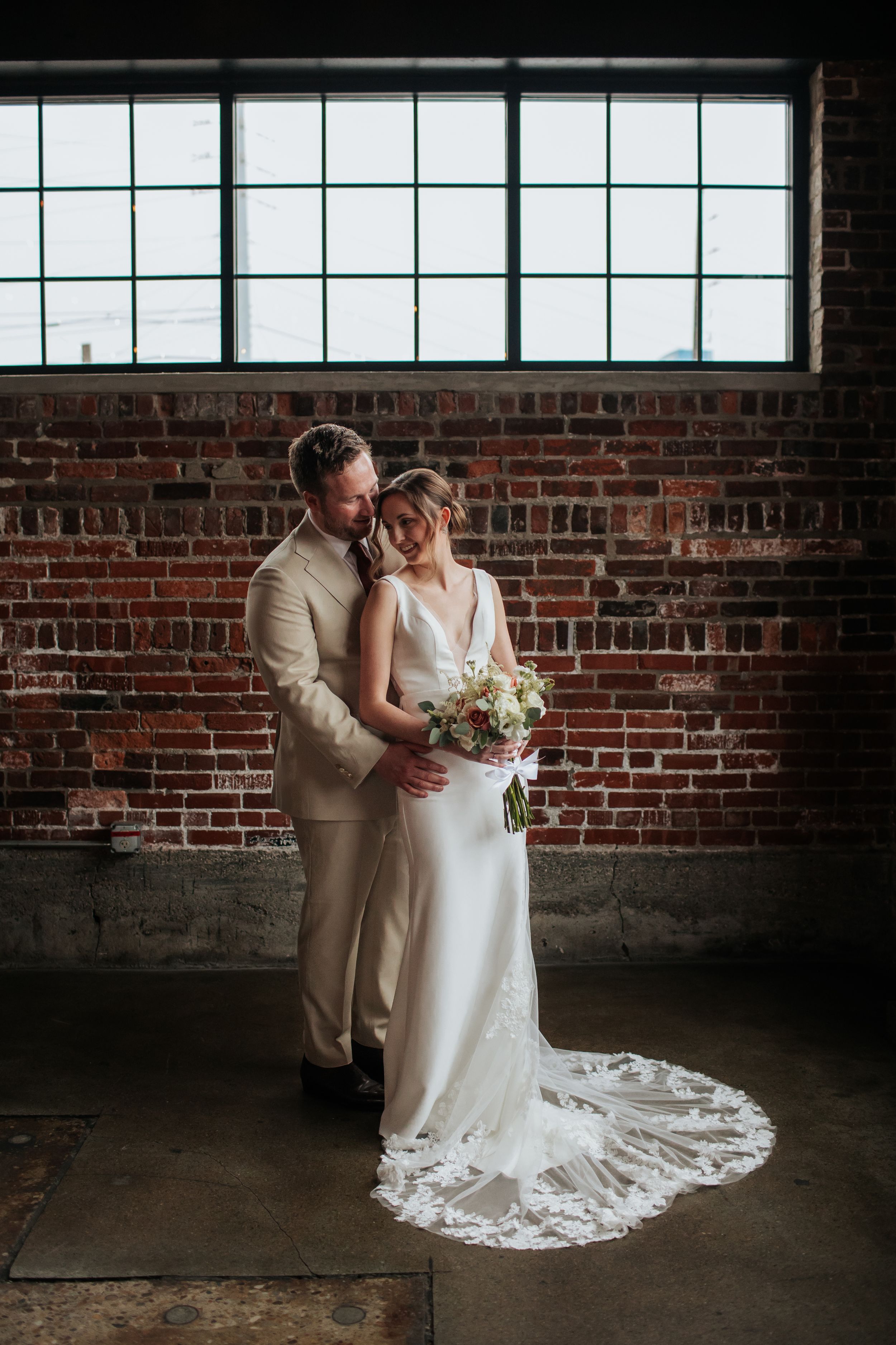 A romantic wedding couple embraces in front of an industrial brick wall with large windows and a flowing lace train.