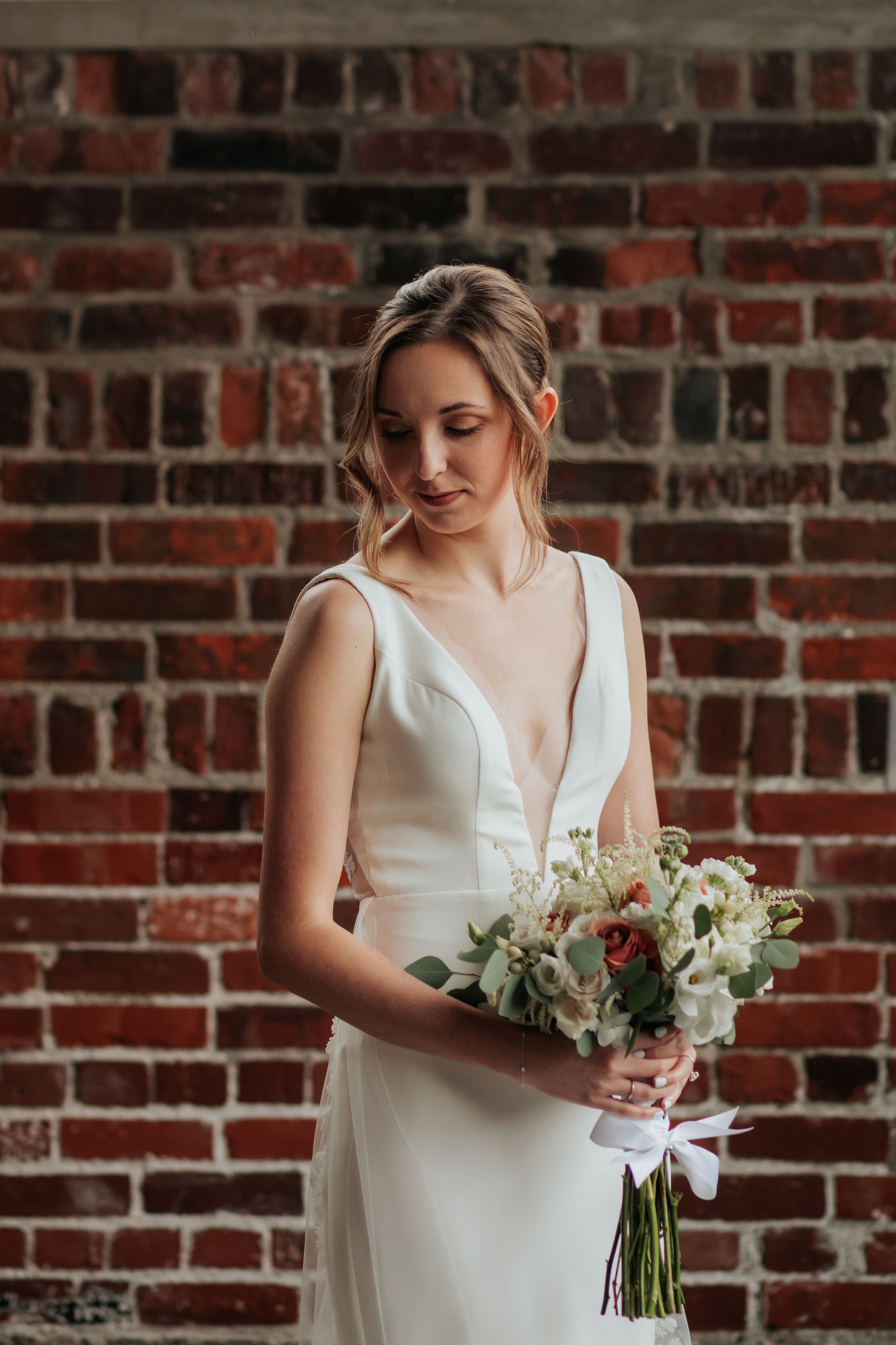 A white bridal bouquet with delicate flowers and greenery is held against a rustic brick wall backdrop.