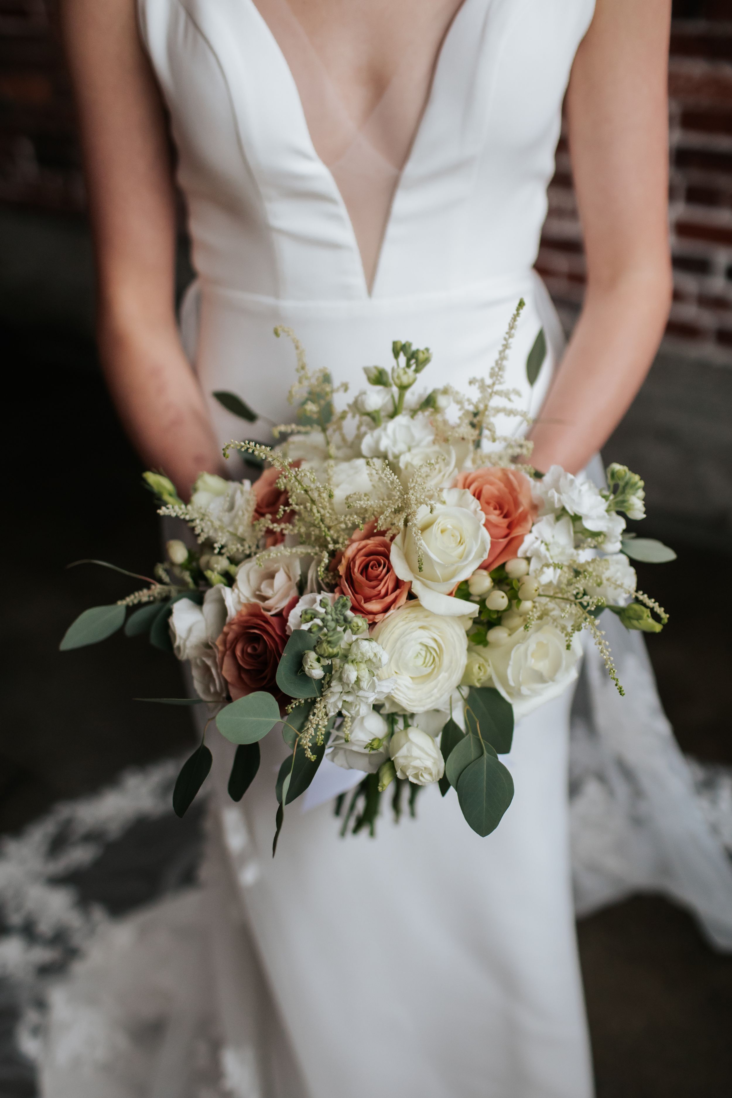 A bride in a white plunging neckline gown holds an elegant bouquet of coral and white roses with eucalyptus greenery.