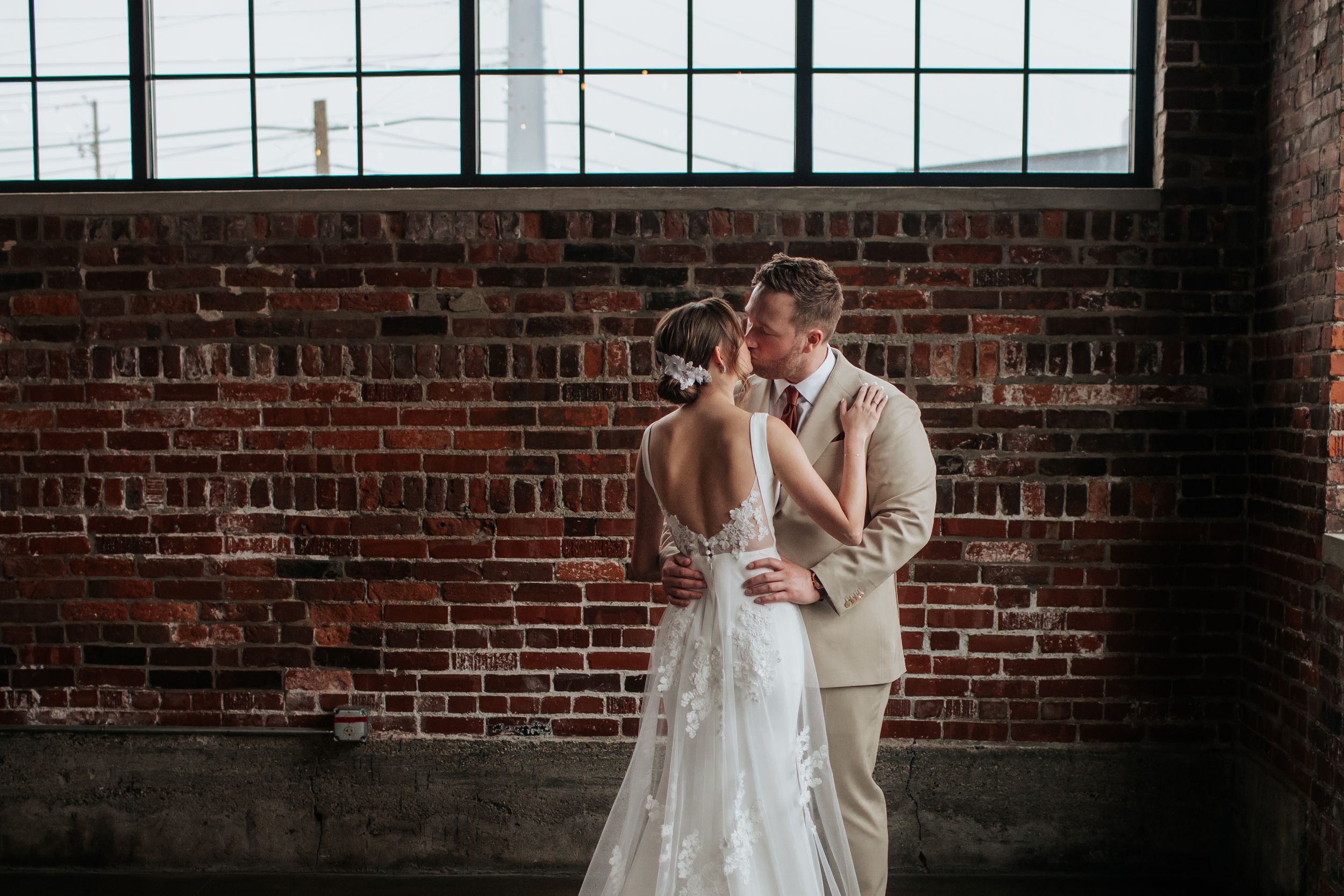 A romantic couple shares a tender moment in front of an industrial brick wall with large windows in a rustic venue.