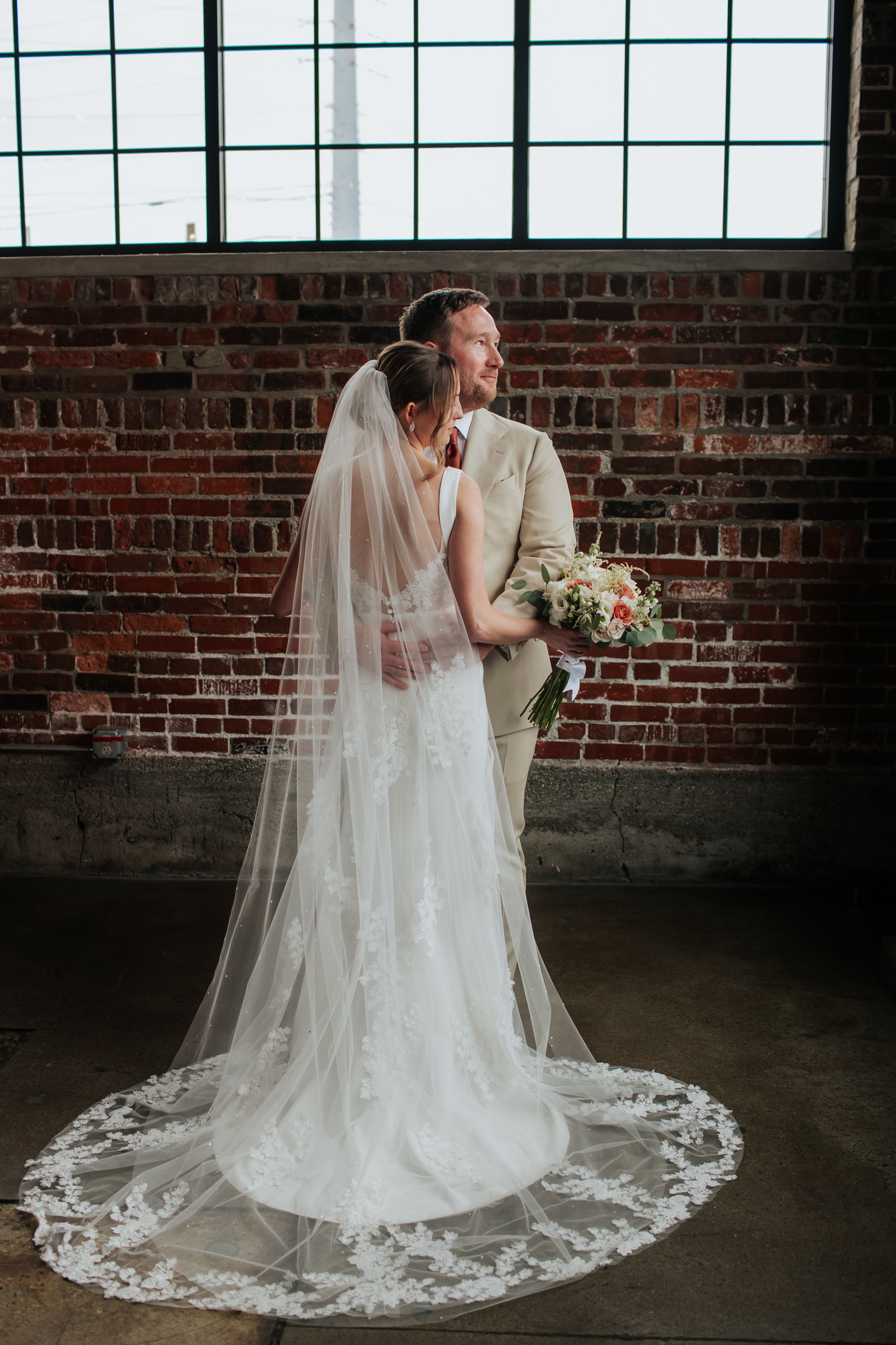 A romantic wedding couple embraces in front of a rustic brick wall as the bride's lace veil trails behind her.