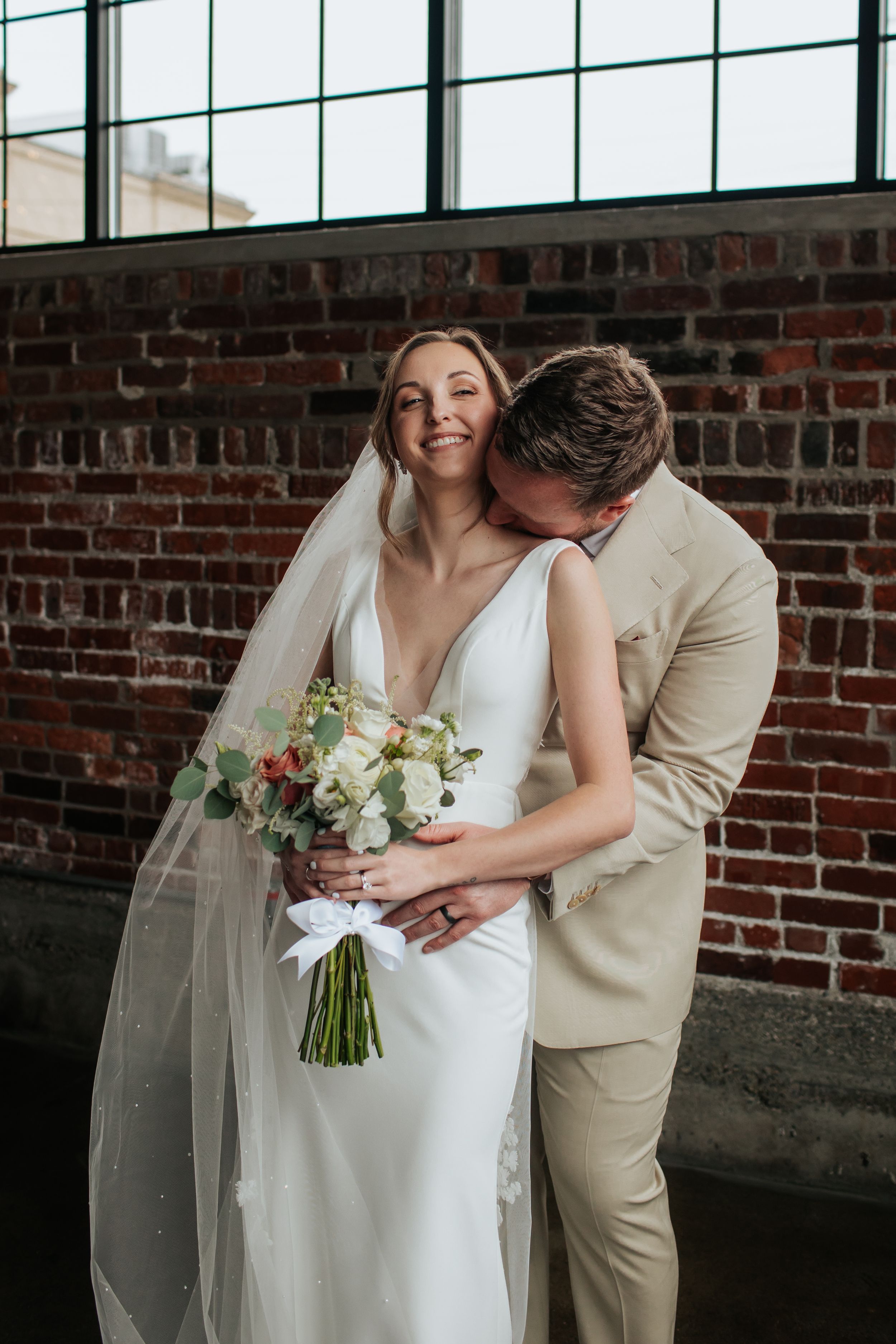 A tender wedding moment captured against a rustic brick wall as a couple embraces with a beautiful white bouquet.