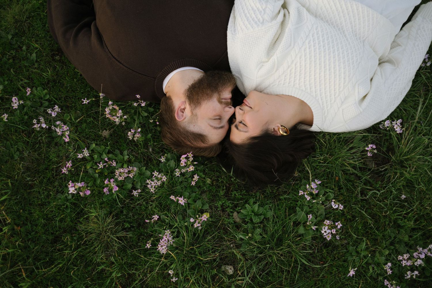 A couple lying in grass with white cherry blossom petals scattered around them during springtime.
