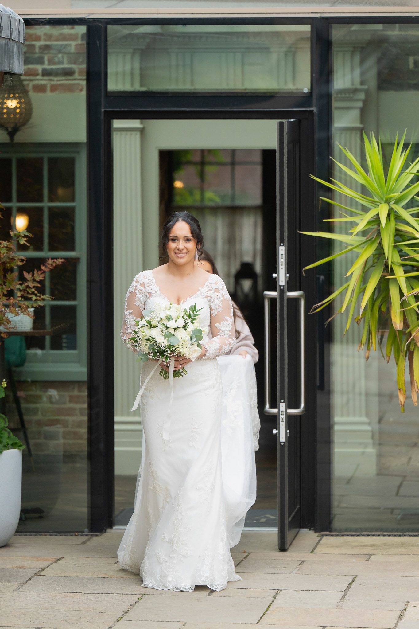 A bride in a white lace wedding dress holds a bouquet while standing in front of a modern building entrance.