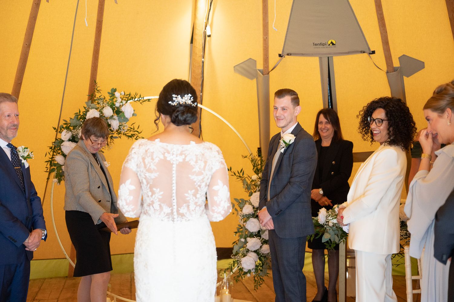 Wedding guests dance and celebrate on a dance floor inside a warmly lit tent decorated with string lights and floral arrangements.