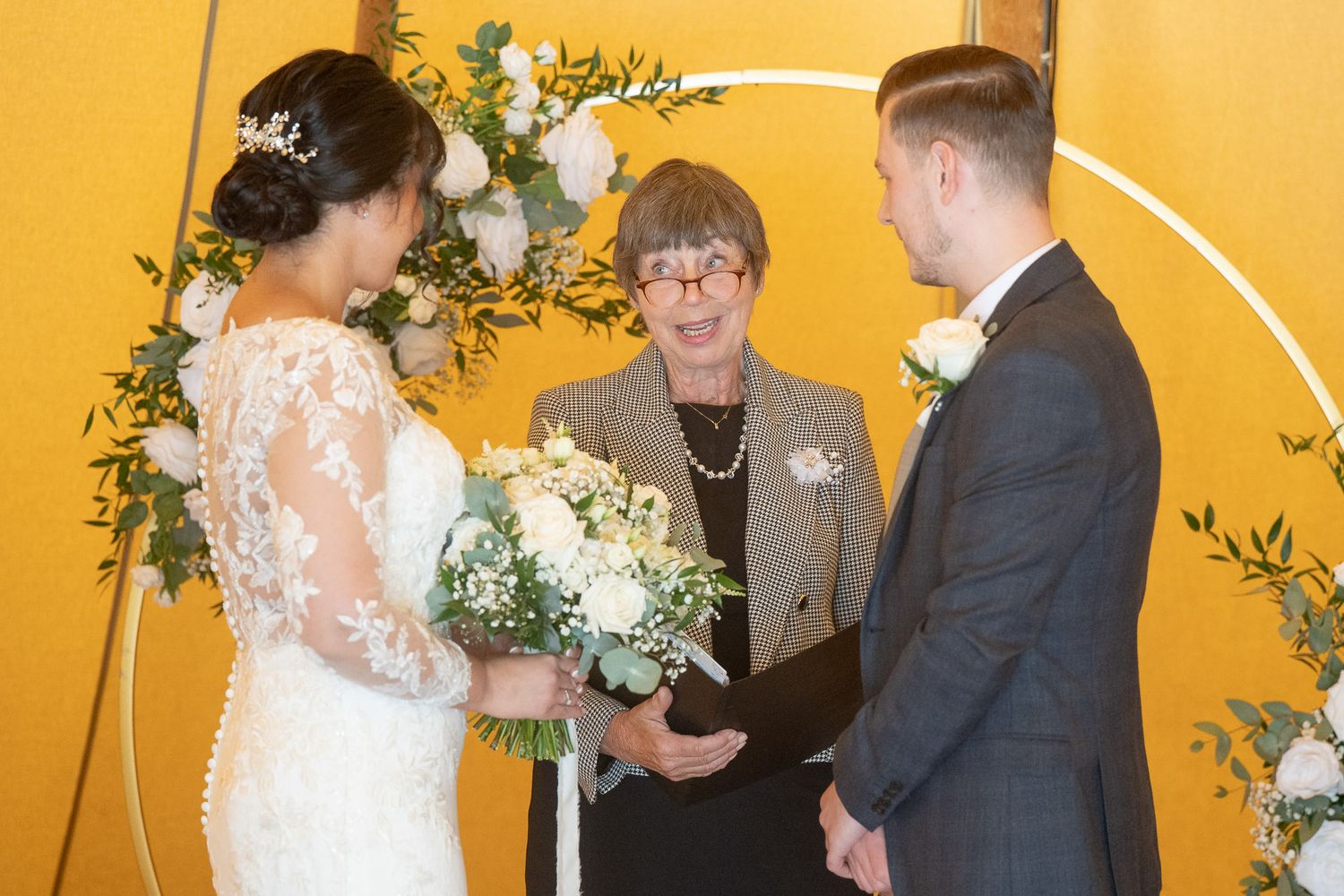 A wedding ceremony takes place in front of a bright yellow wall decorated with green foliage.
