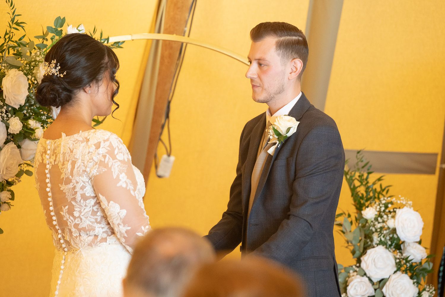 A couple shares an intimate moment during their wedding ceremony in front of a yellow wall with floral decorations.