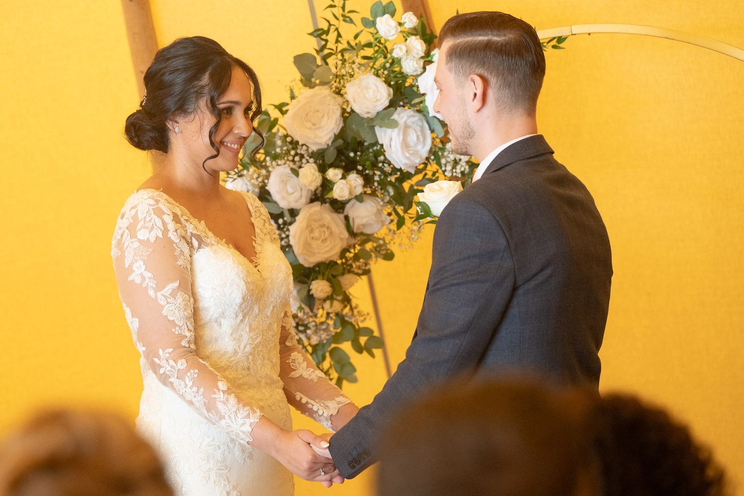 A couple stands at the altar during their wedding ceremony in front of a yellow wall with floral arrangements.