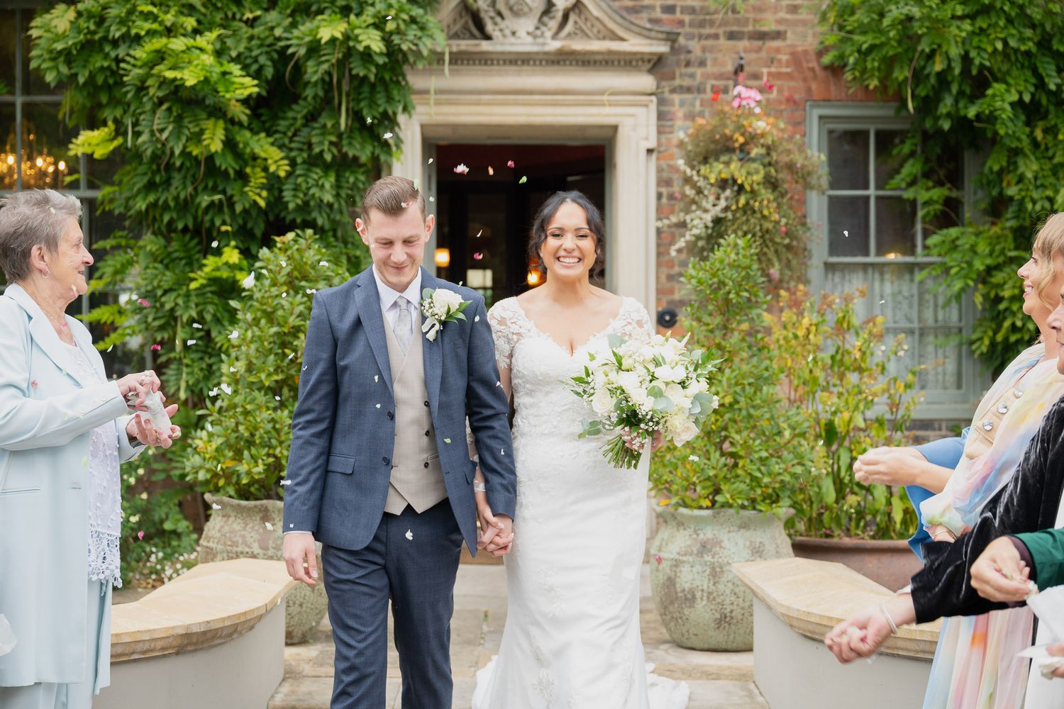 Wedding party celebrates in front of historic brick building with lush green foliage.