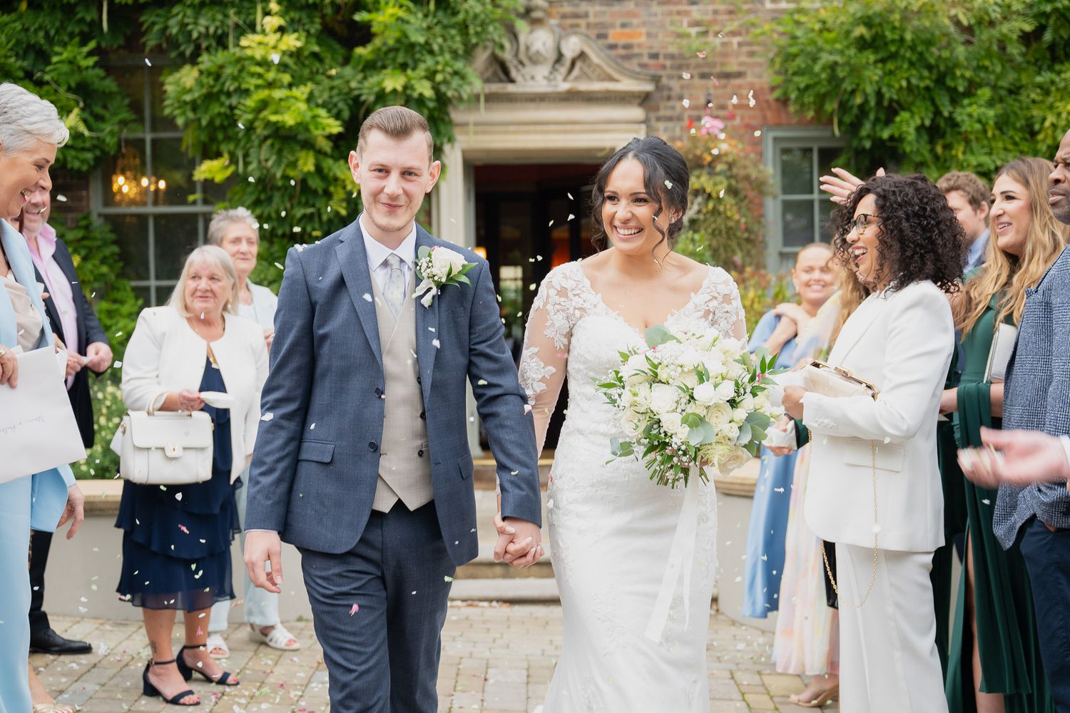 Wedding guests celebrate joyfully in an outdoor garden setting with brick walls and lush greenery in the background.