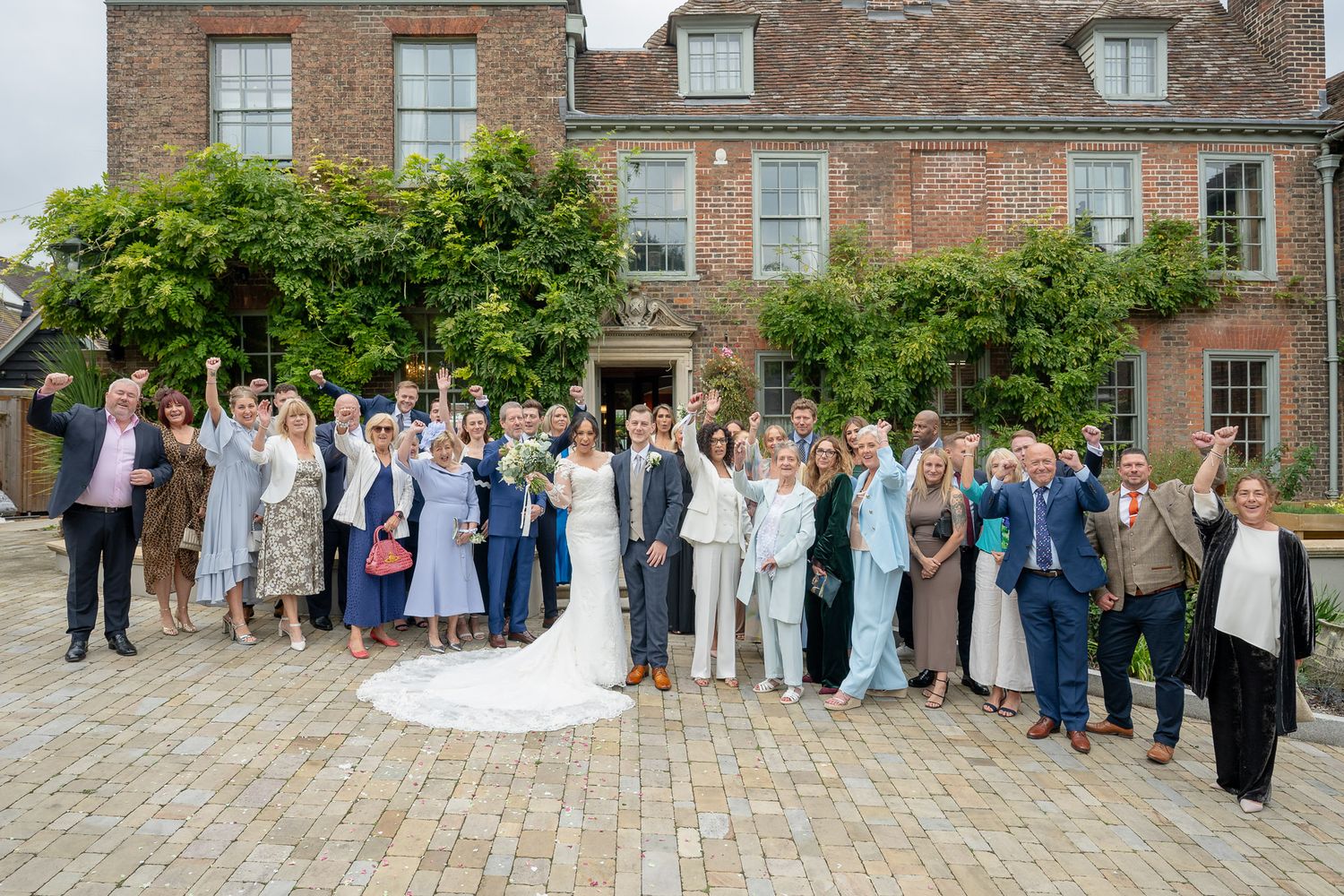 Wedding party guests gather on a historic brick courtyard for formal group photos on a sunny day.